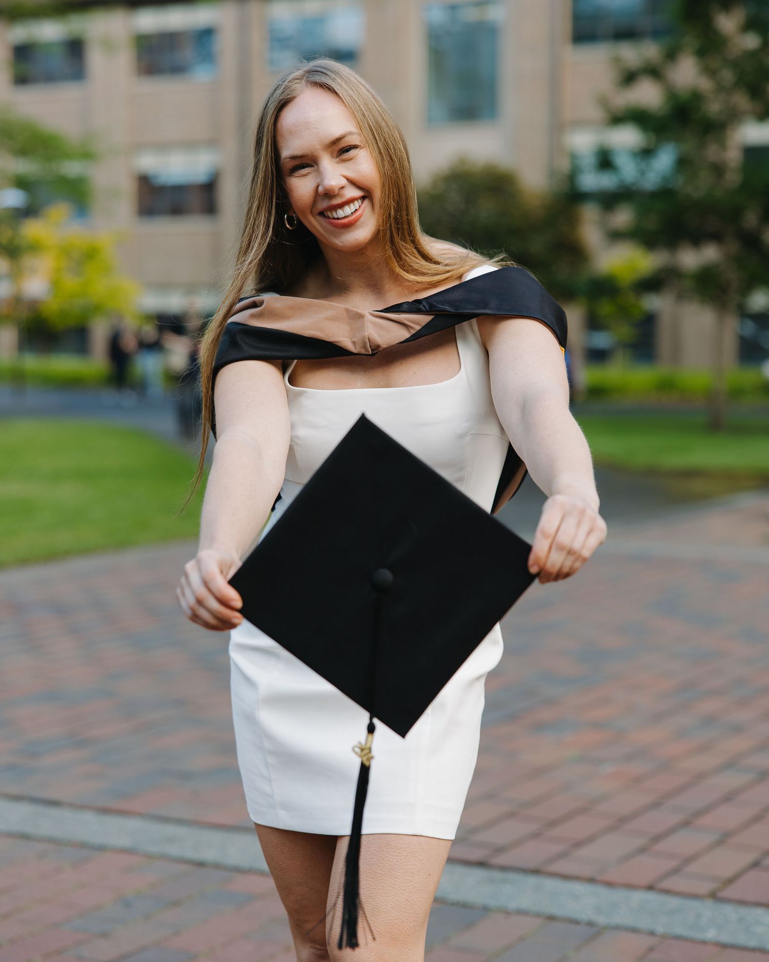 Joyful graduate in white dress holding black mortarboard cap on campus.