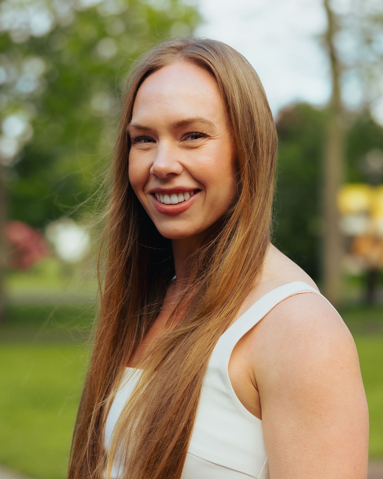 Smiling woman with long blonde hair wearing a white sleeveless top in a park.
