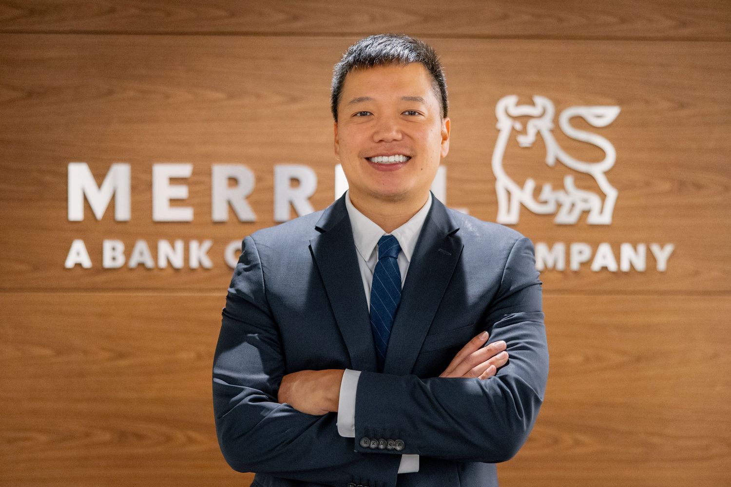 Professional Asian man in dark suit standing confidently with arms crossed in front of Merrill Lynch sign.
