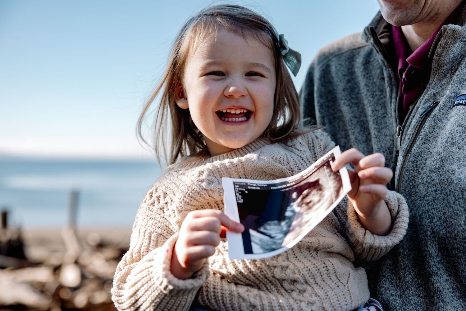 Laughing toddler girl holds ultrasound photo at beach.