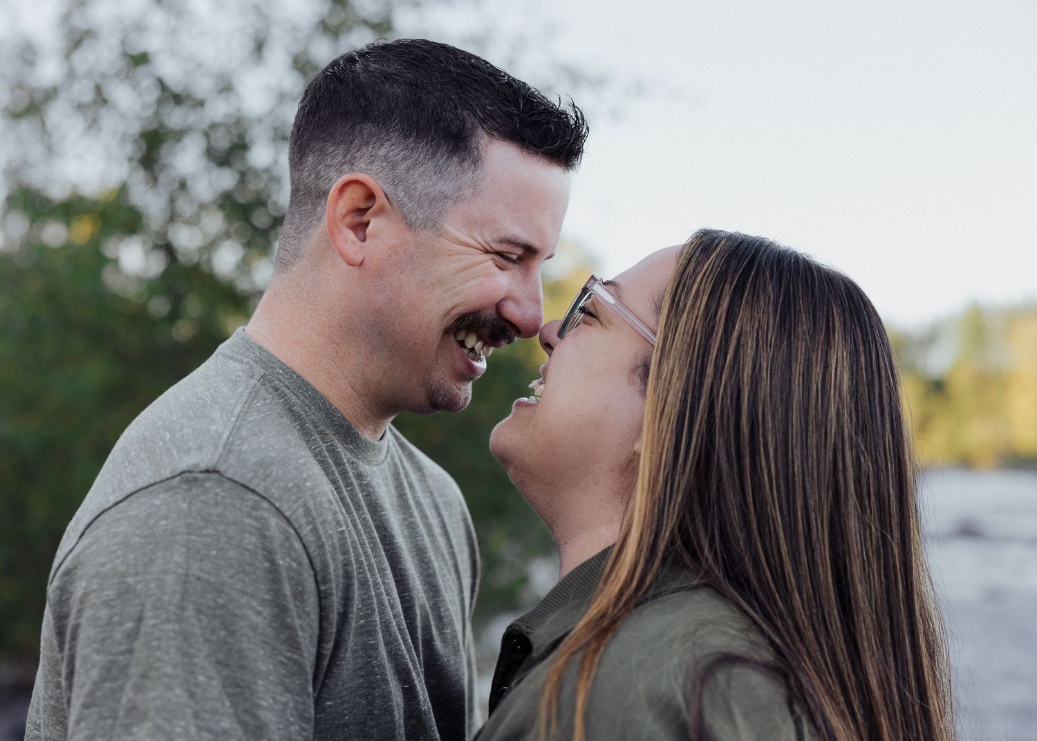 Laughing couple facing each other closely outdoors near water, sharing a playful and joyful romantic moment.