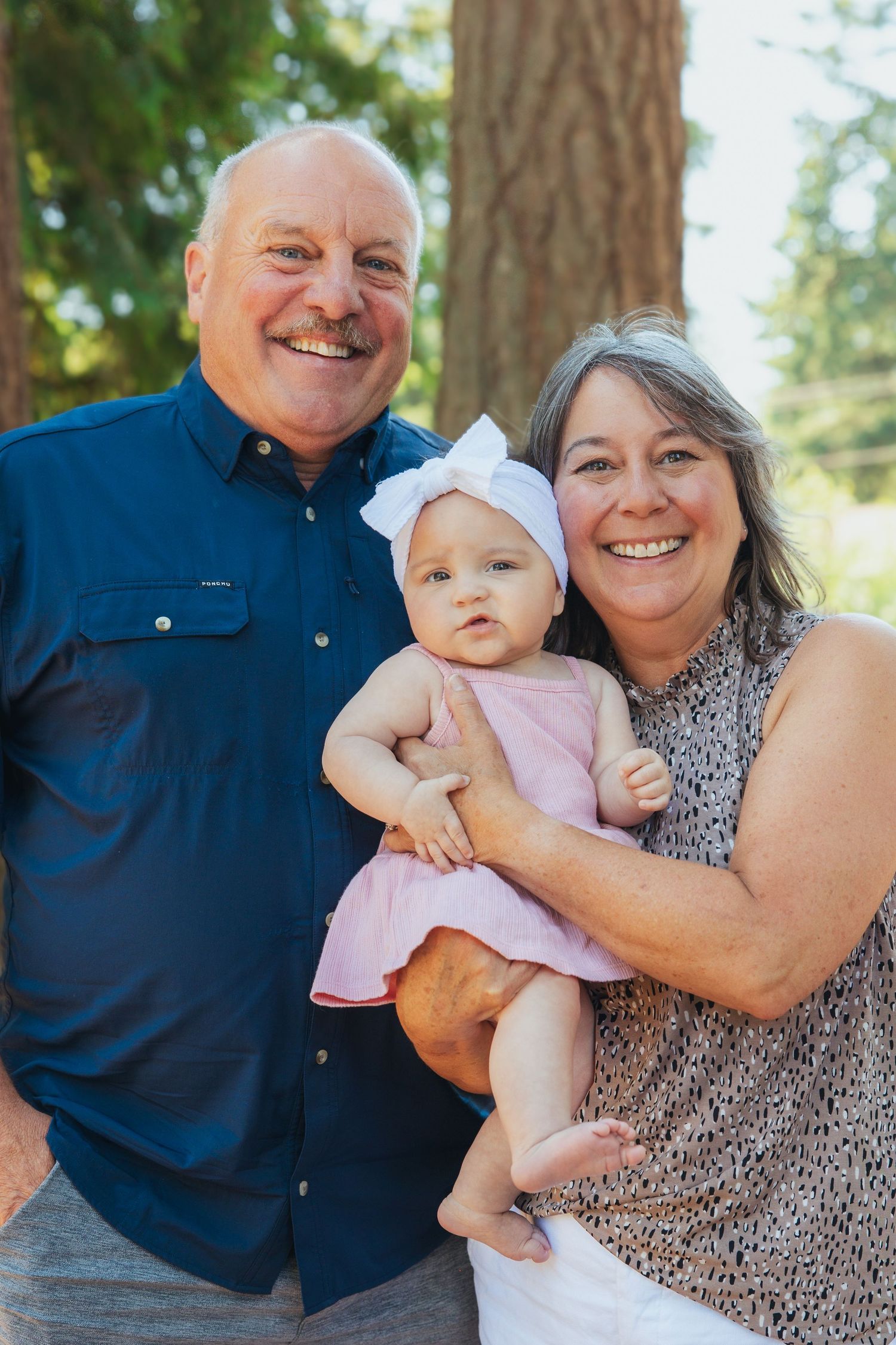 Grandparents smiling with a baby girl in a pink dress outdoors.