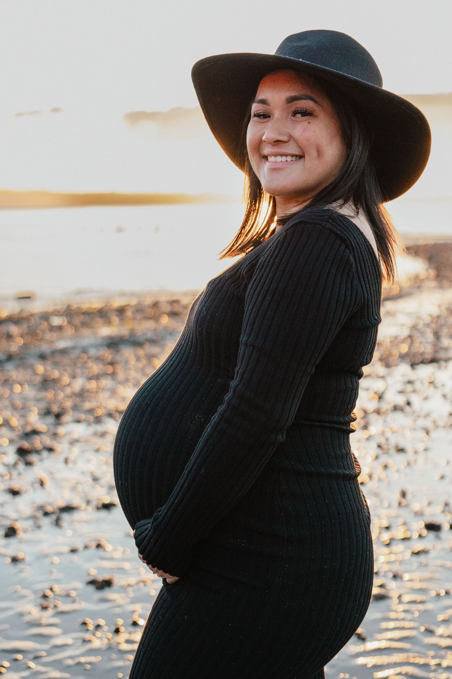 Smiling pregnant woman in black dress and hat holding belly on a beach at golden sunset.