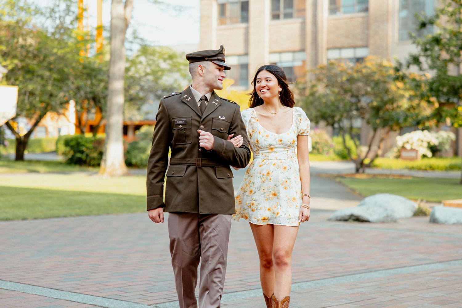 Couple walking together on campus plaza, man in Army dress uniform and woman in white floral sundress smiling.