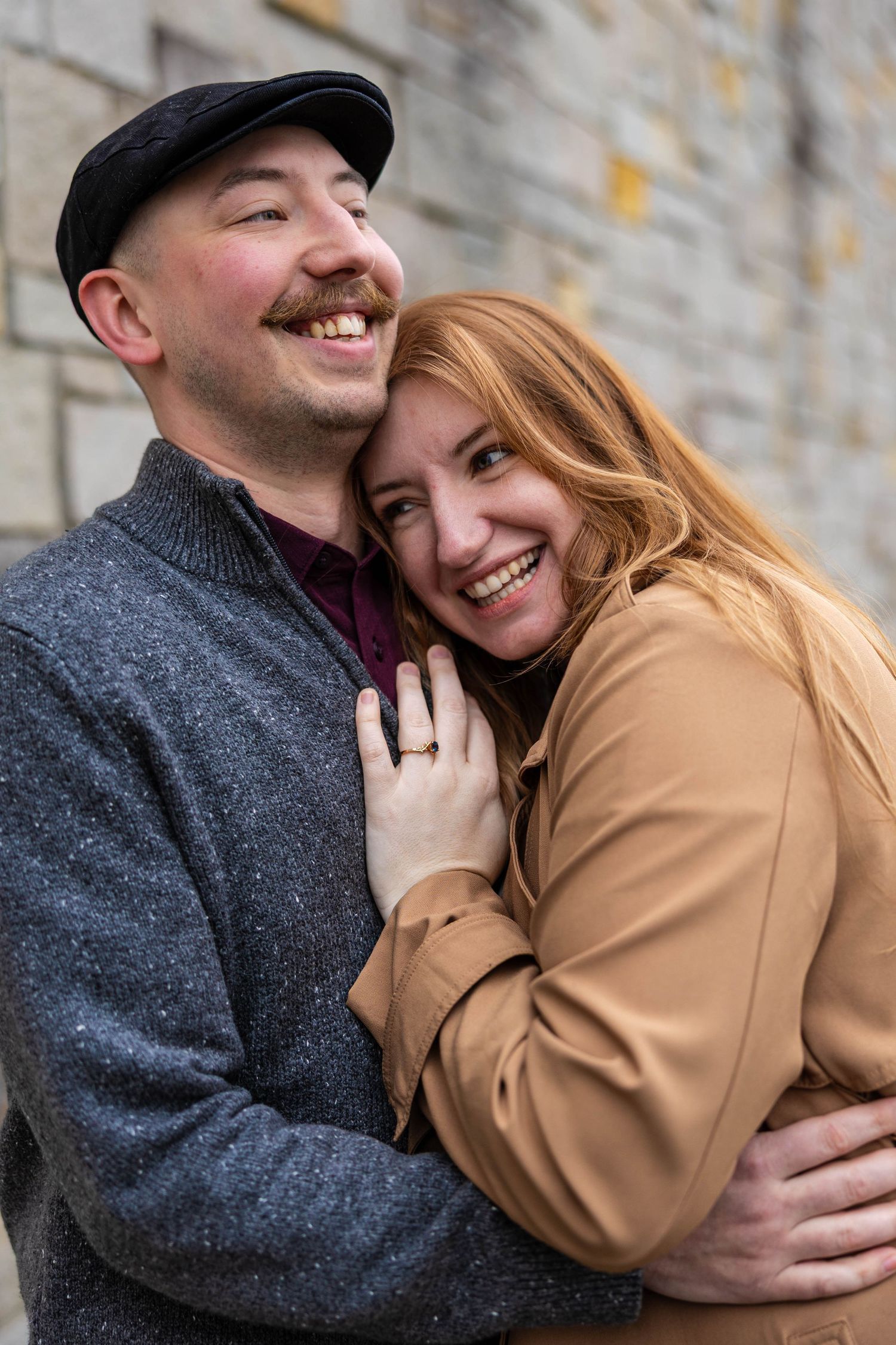 Smiling couple embracing against a stone wall, woman with red hair wearing engagement ring in a warm beige coat.