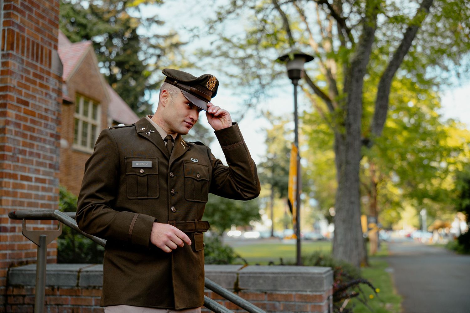 A young man in military dress uniform tips his hat while standing on steps near a brick building outdoors.