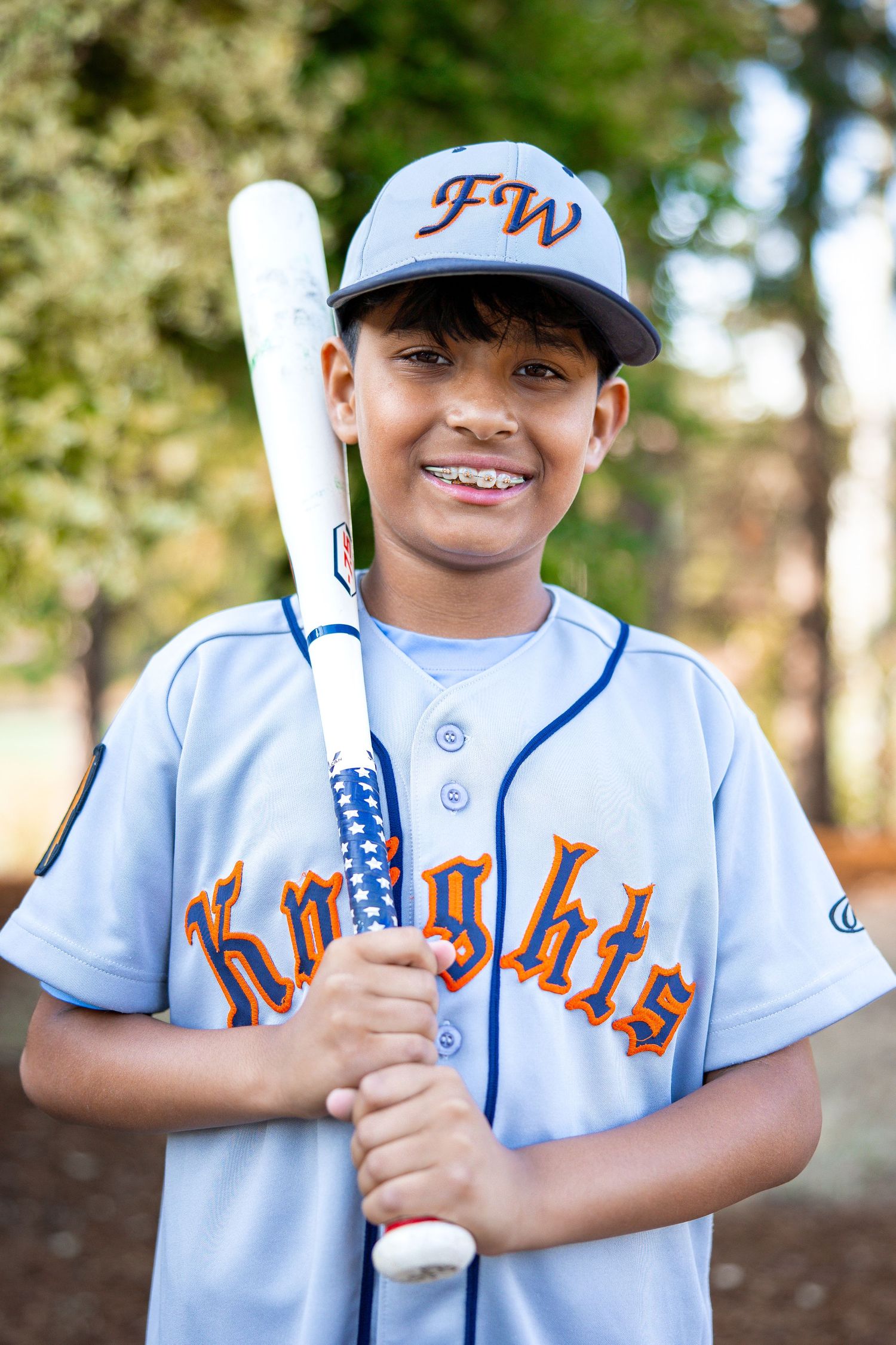 Smiling young baseball player in gray FW Knights uniform holds a bat for an outdoor portrait.