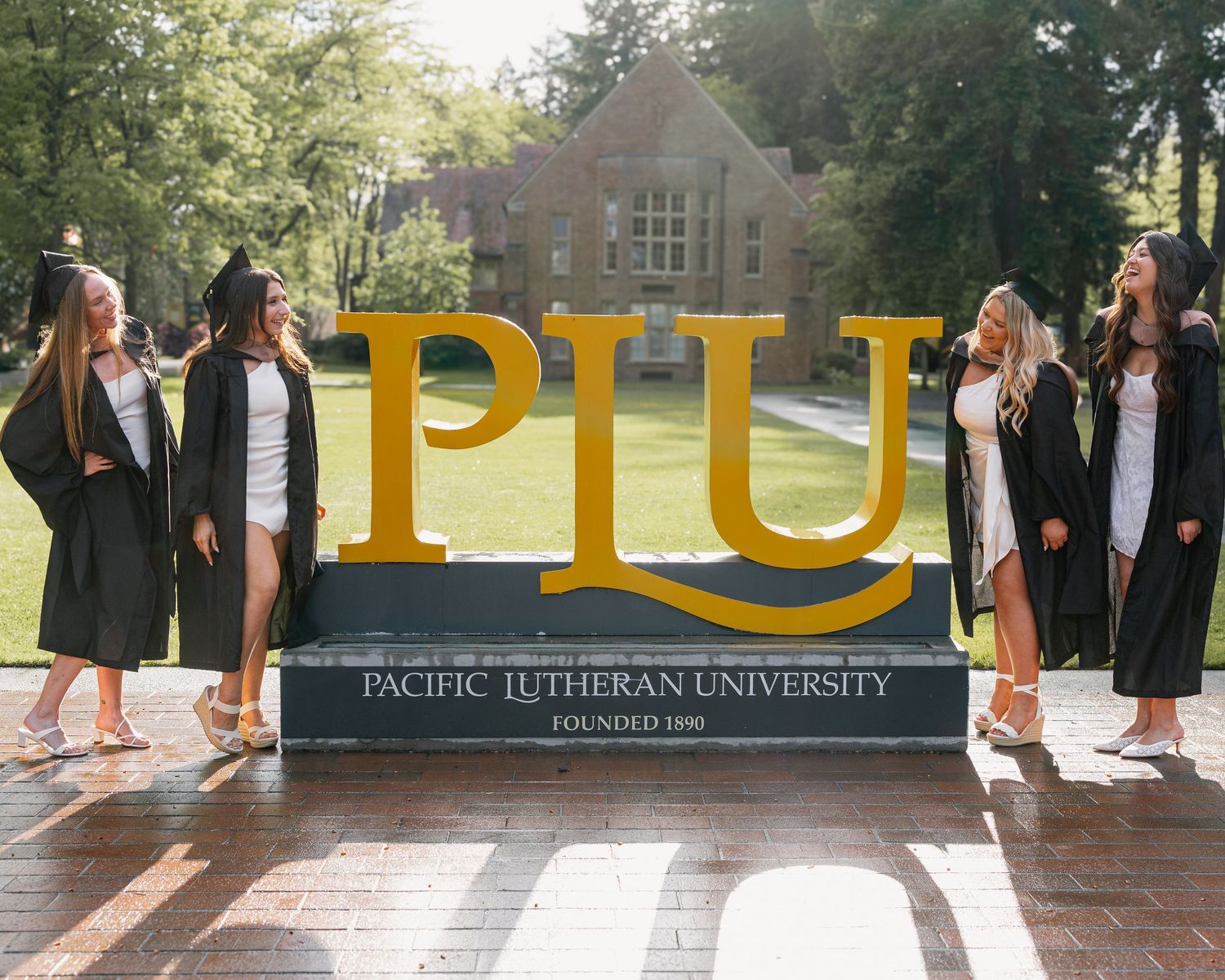 Four female graduates in black robes posing beside Pacific Lutheran University PLU campus sign.