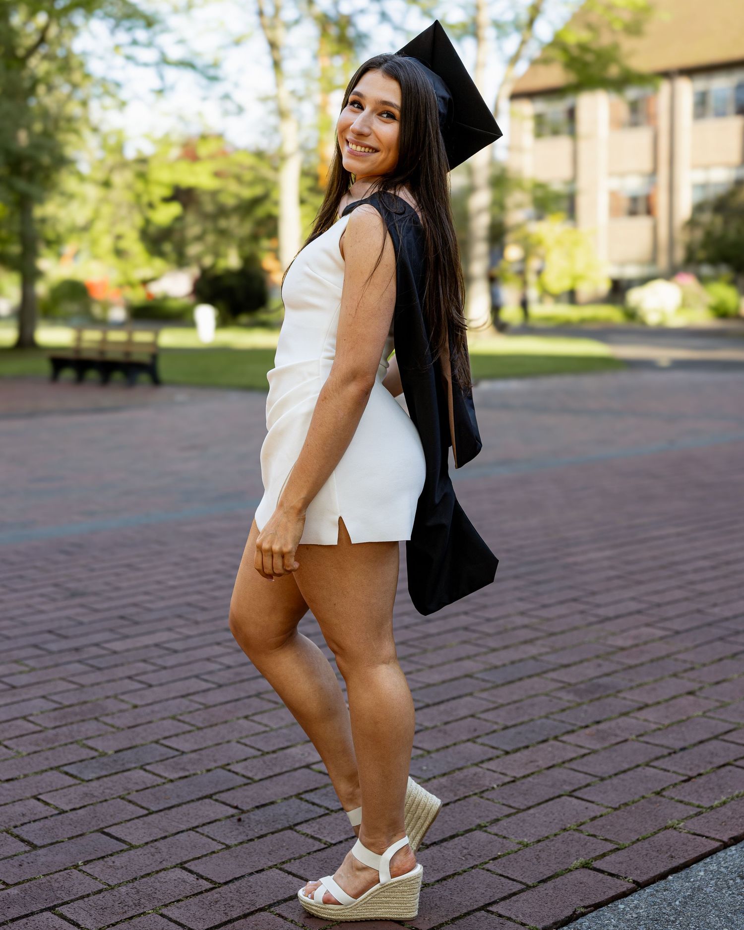 Graduate in white mini dress and wedge heels posing on brick campus walkway.