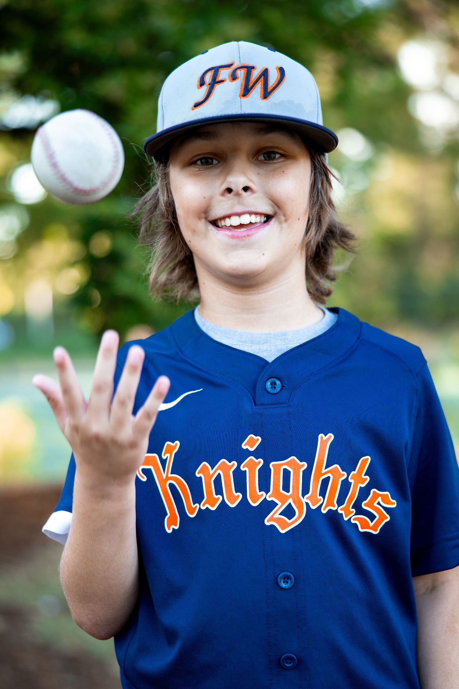 Smiling young boy in navy FW Knights baseball jersey tosses a baseball into the air outdoors.