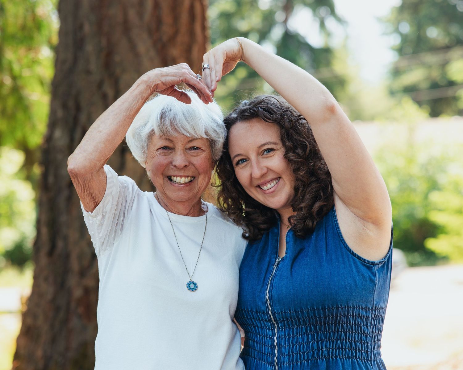 Two women smile joyfully while forming a heart shape with their hands overhead in a sunny outdoor setting.