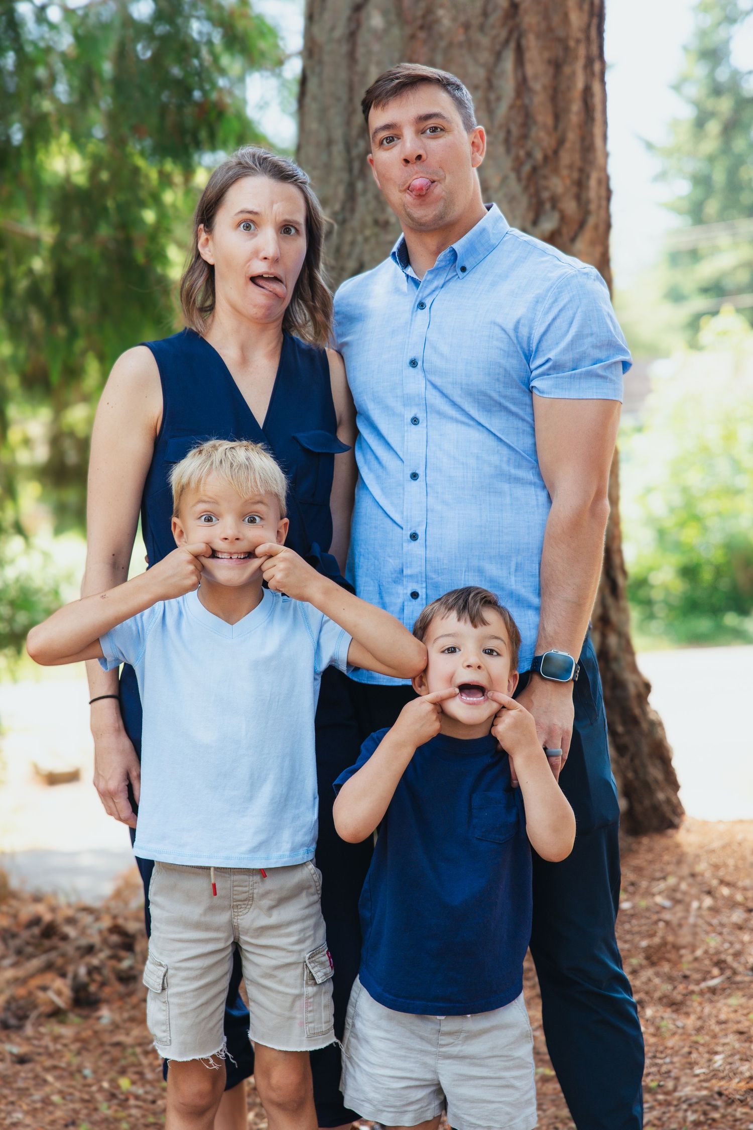 Family of four making funny faces together outdoors near a large tree.