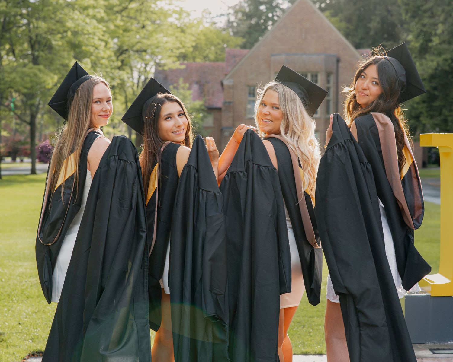Four female graduates in black robes showing colorful hood linings while posing on campus lawn.