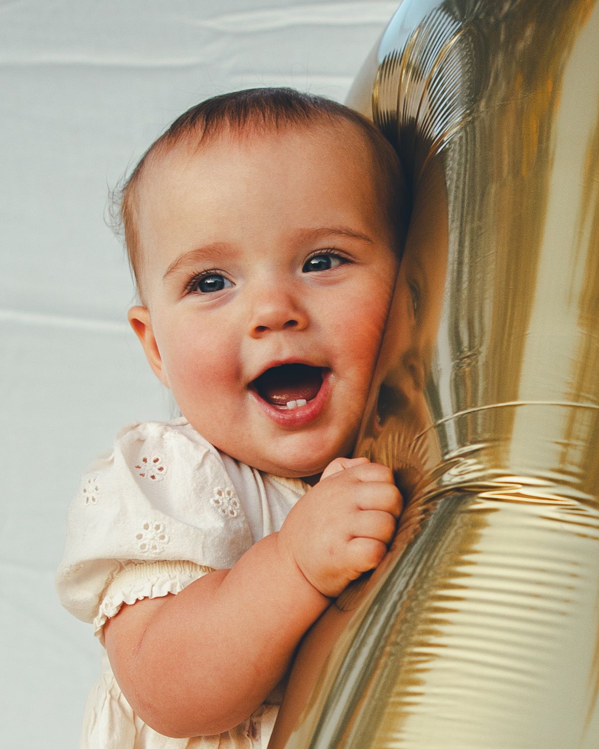 Happy laughing baby in white dress hugging a large gold balloon.