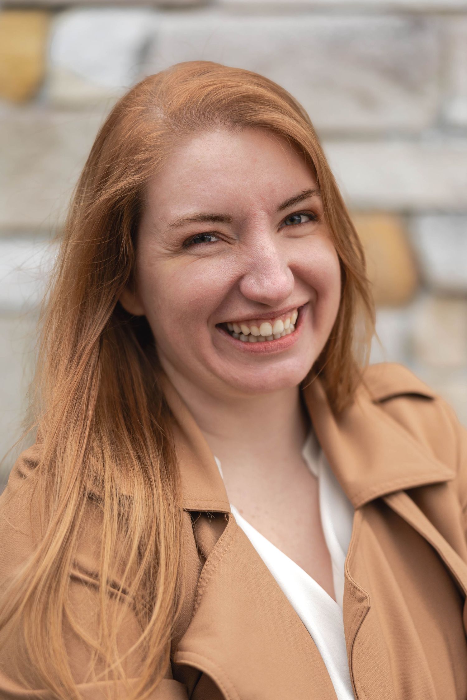 Smiling young woman with red hair wearing a tan jacket near a stone wall.