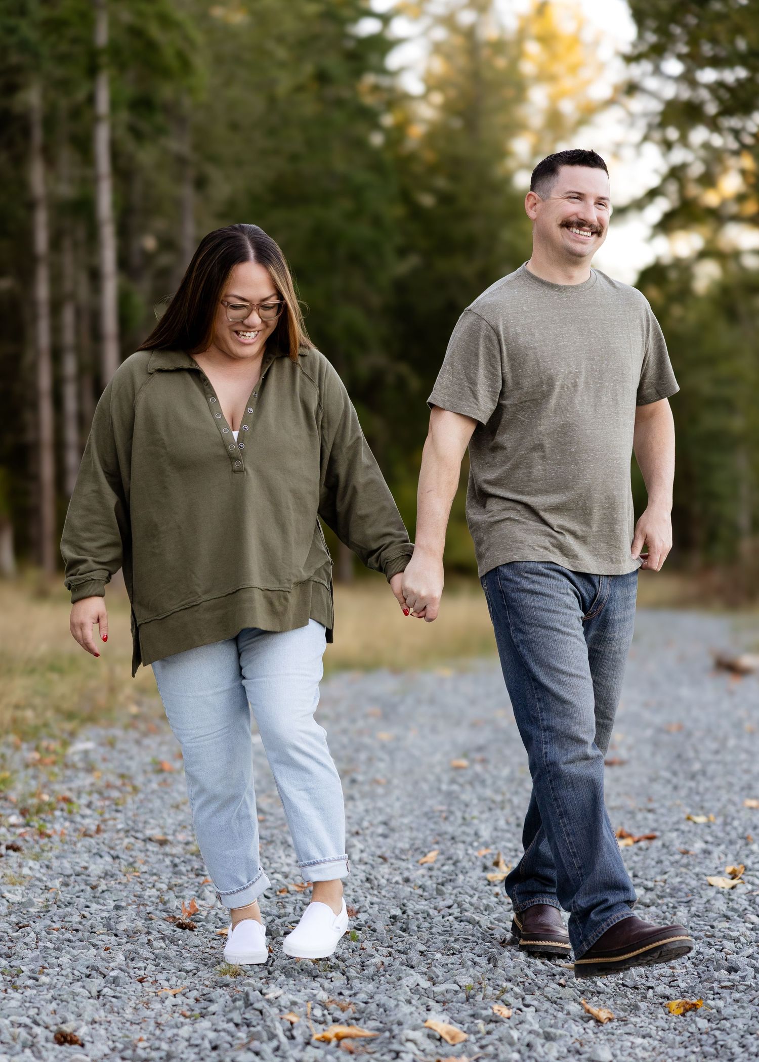 Engaged couple holding hands and laughing while walking on a gravel path in nature.