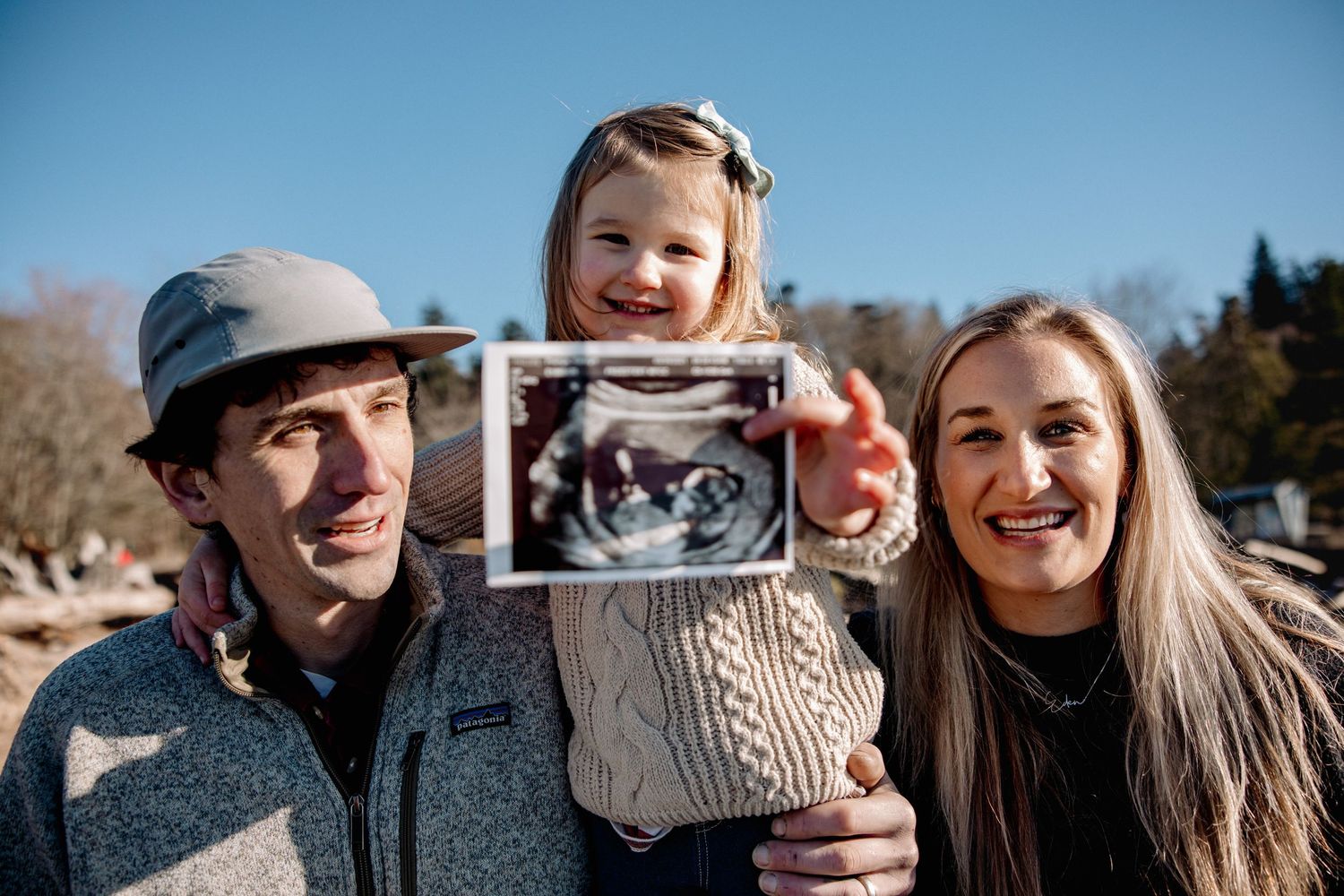Smiling family holds ultrasound photo for pregnancy announcement outdoors.