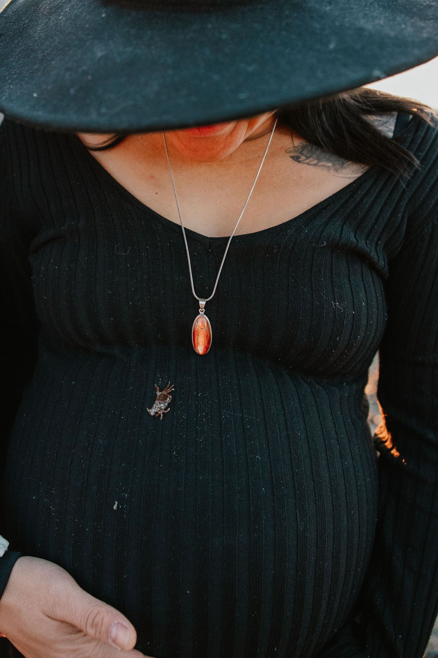 Pregnant woman in black ribbed dress and wide-brim hat wearing a silver pendant necklace with orange stone.