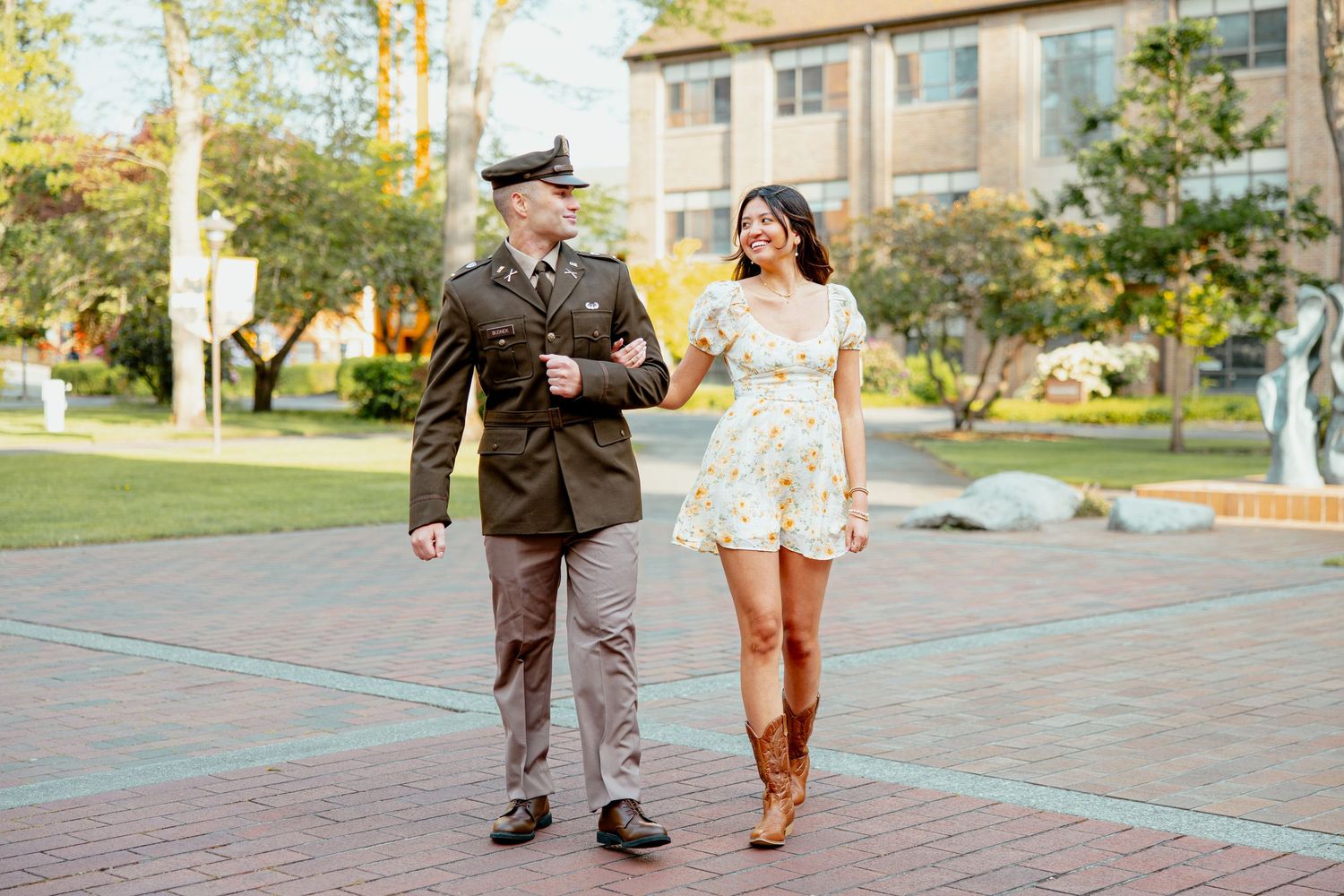Military man in uniform walking with woman in floral dress on a sunny campus pathway.