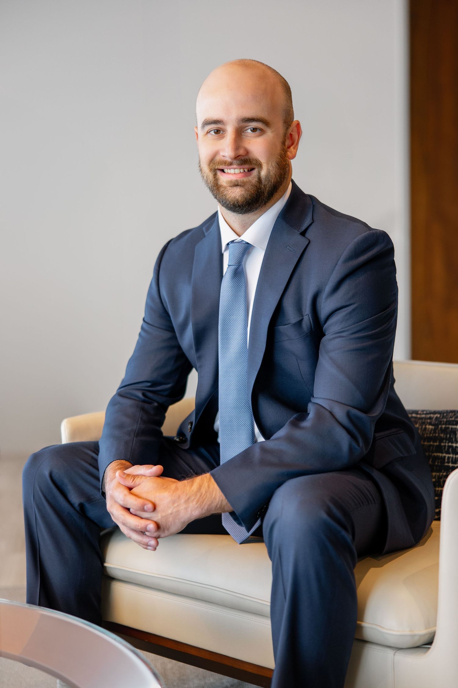 Bald bearded man in navy suit and blue tie seated smiling with hands clasped in modern office.