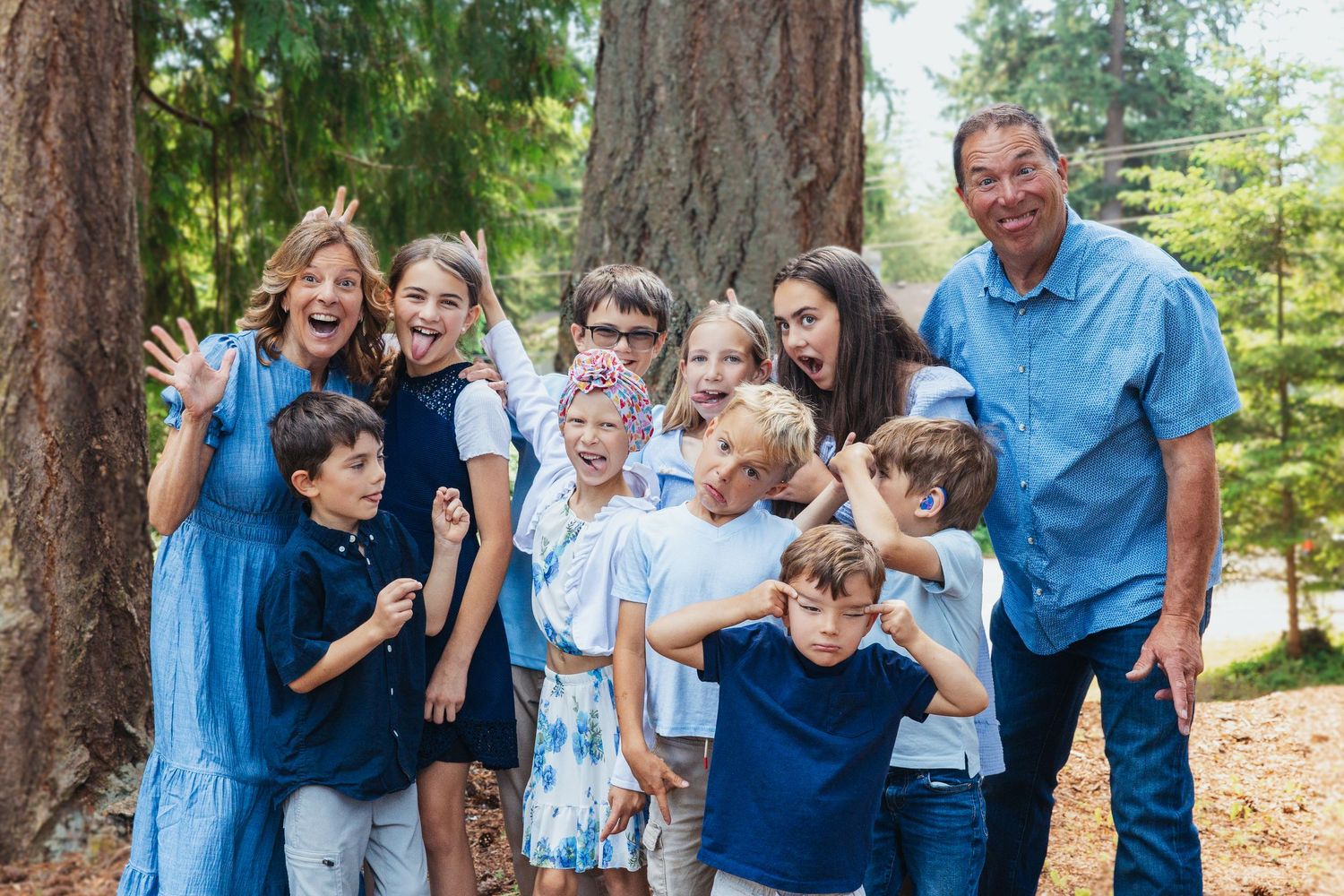 Large joyful family group making silly faces together outdoors near tall trees.