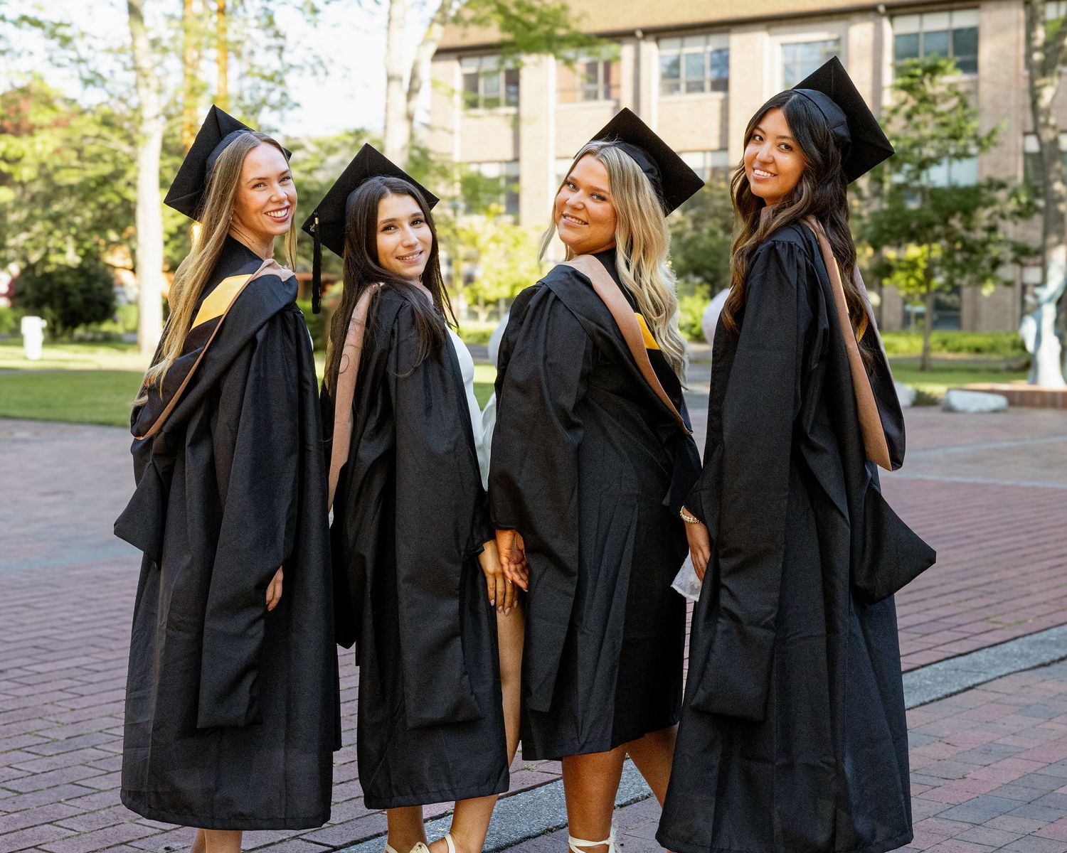 Four female graduates in black caps and gowns pose together outdoors on a brick walkway on a college campus.