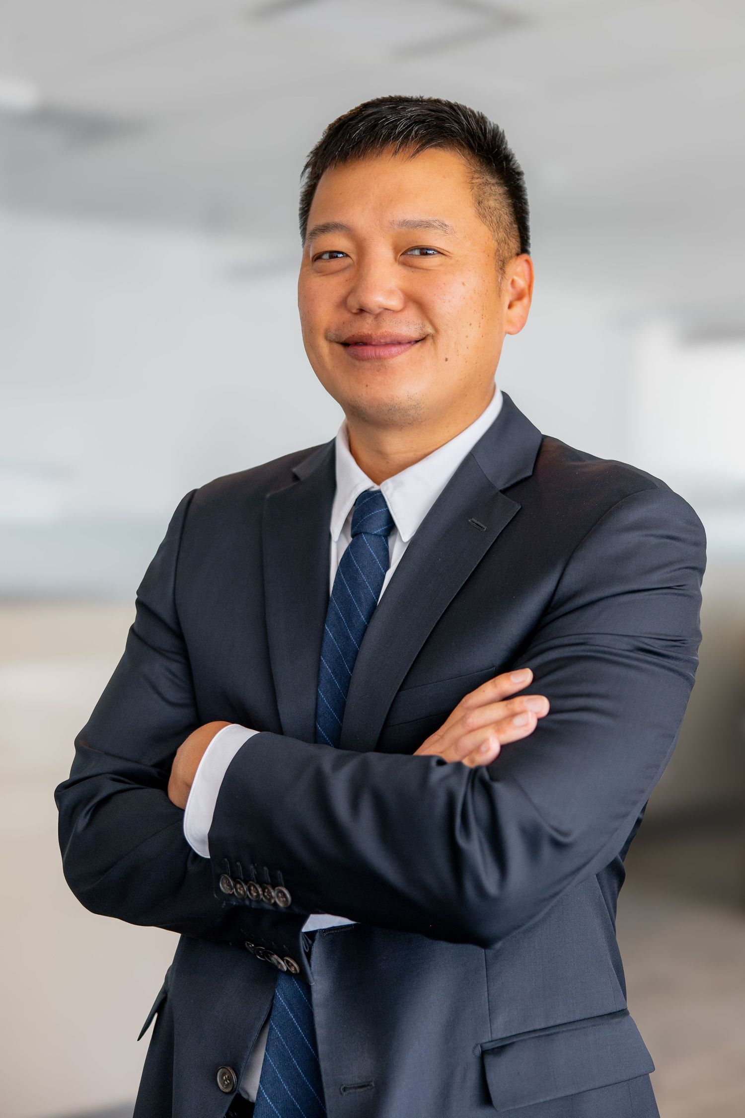 Professional Asian man in navy suit and tie smiling with arms crossed in office setting.