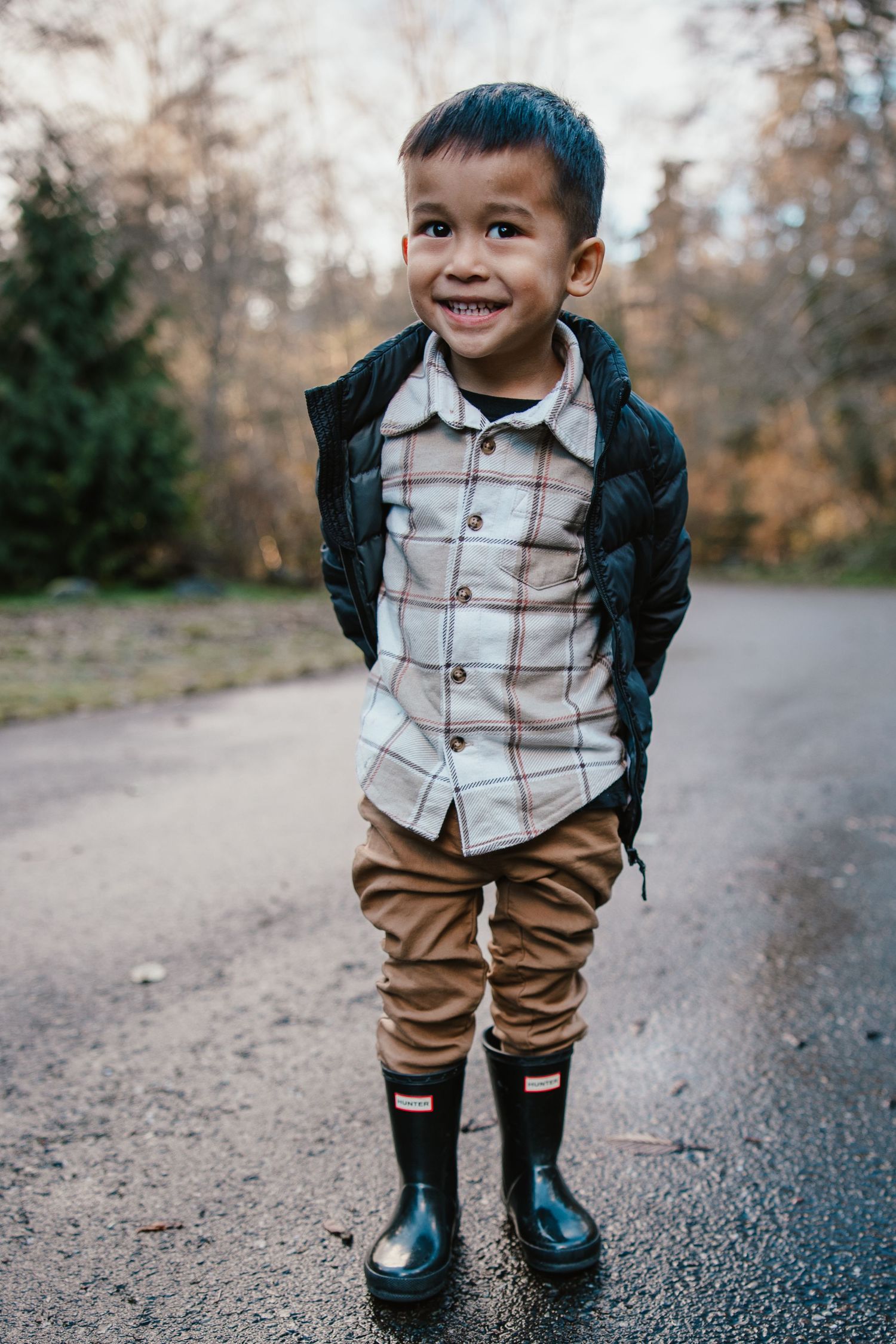 A smiling toddler boy wearing rain boots, plaid shirt, and vest stands on a path surrounded by autumn trees.