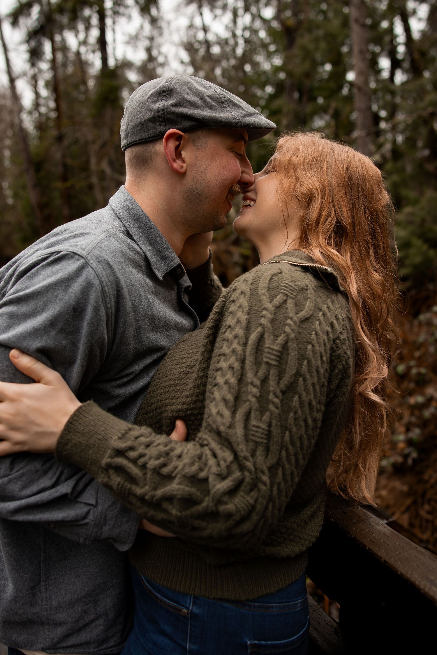 Couple sharing a romantic almost-kiss on a wooden bridge surrounded by evergreen trees.