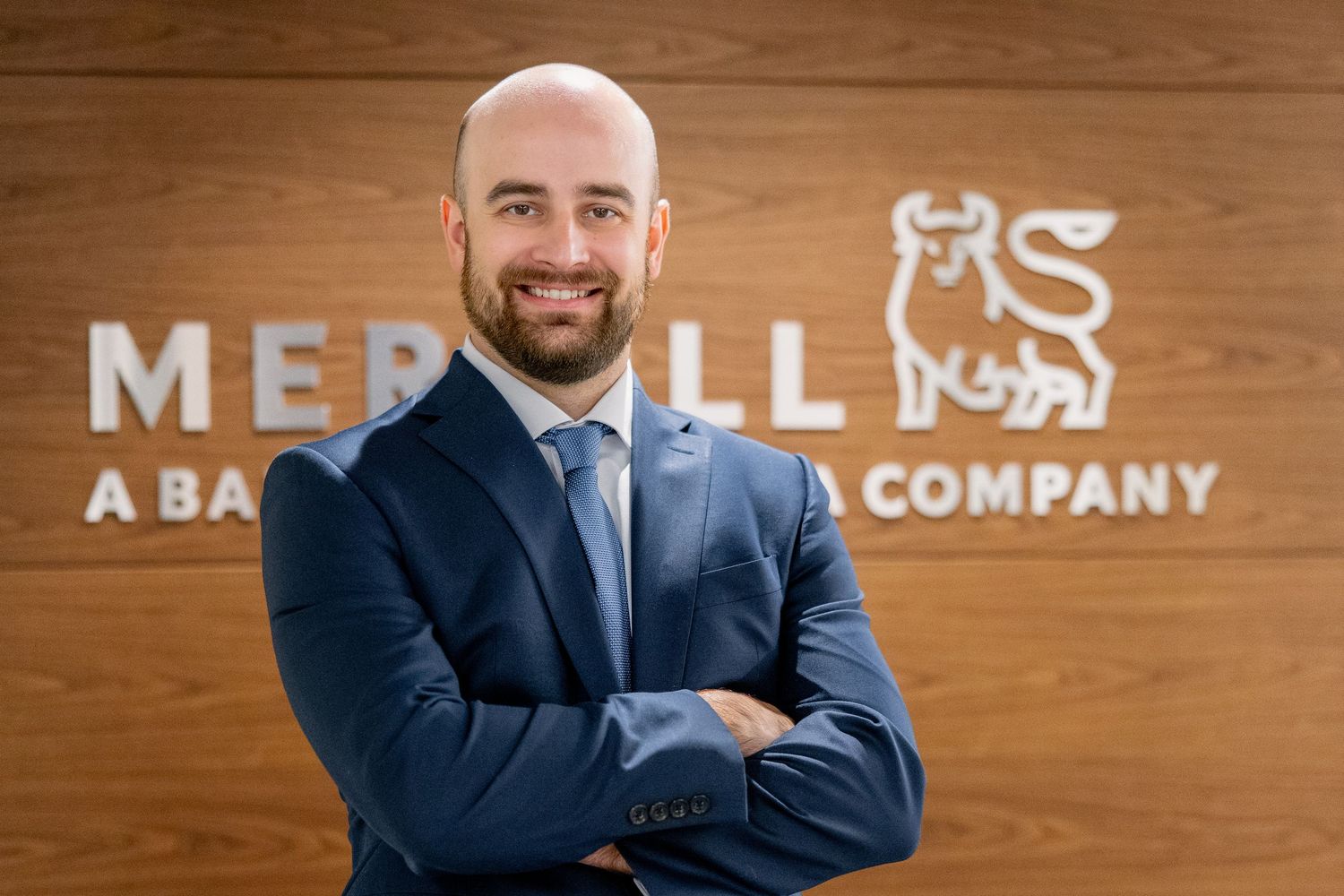 Bald bearded professional in navy suit standing with arms crossed in front of Merrill Lynch logo wall.