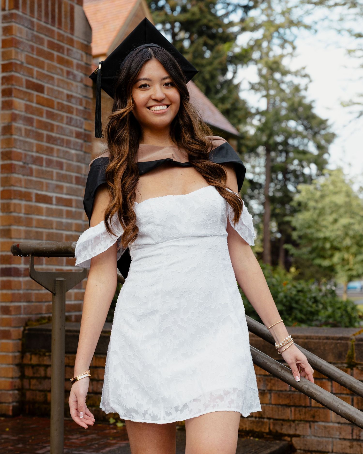 Smiling female graduate in white off-shoulder dress and black graduation cap poses on brick steps near building.