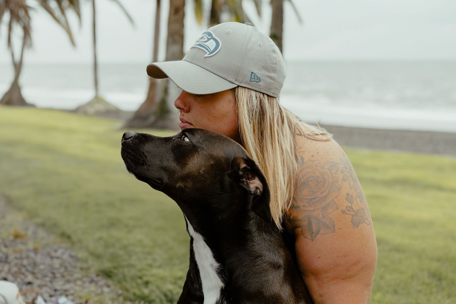 A blonde woman with tattoos and a gray cap shares a tender moment kissing her black dog near the ocean.