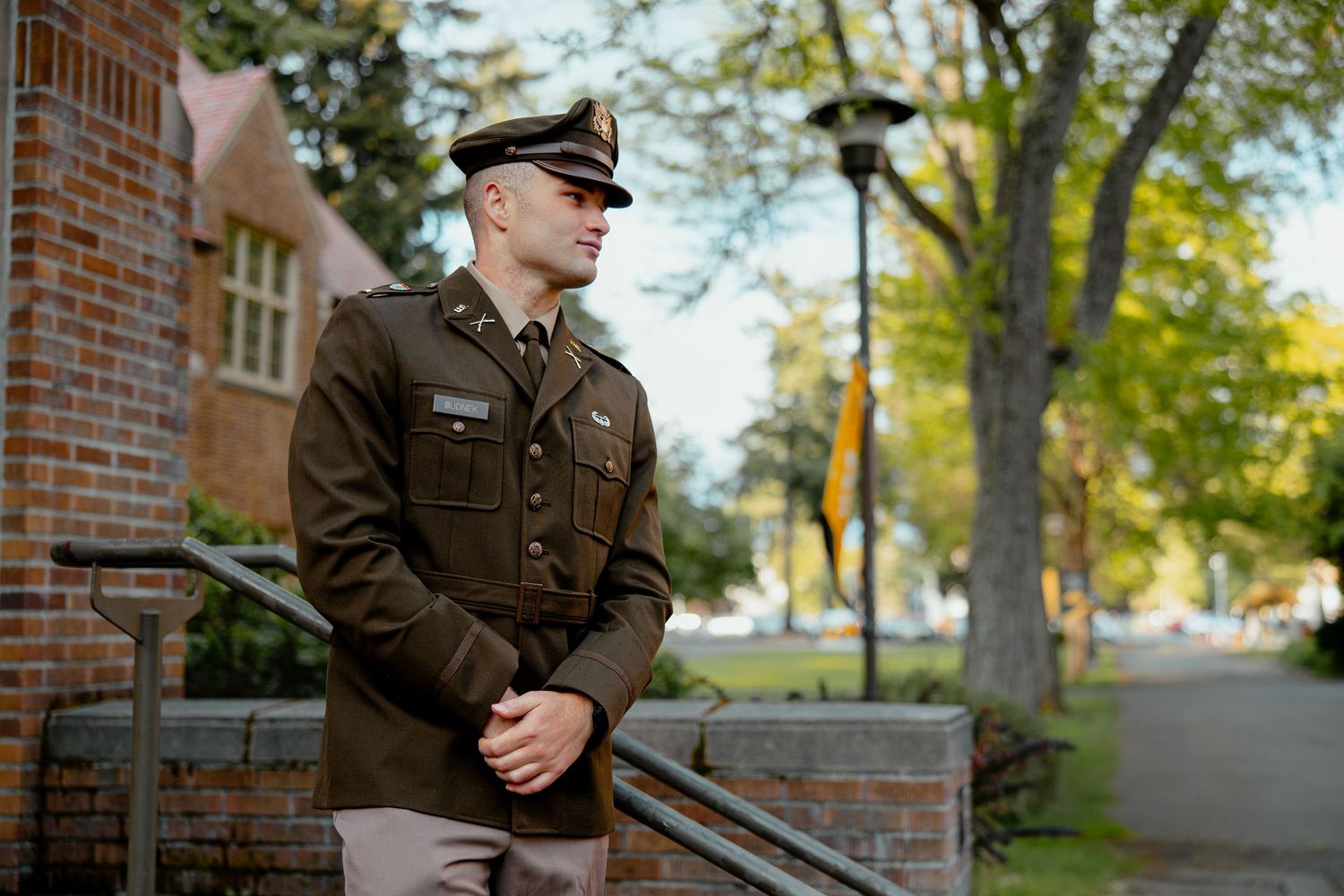 Military officer in dress uniform standing outdoors near brick building and tree-lined path on sunny day.