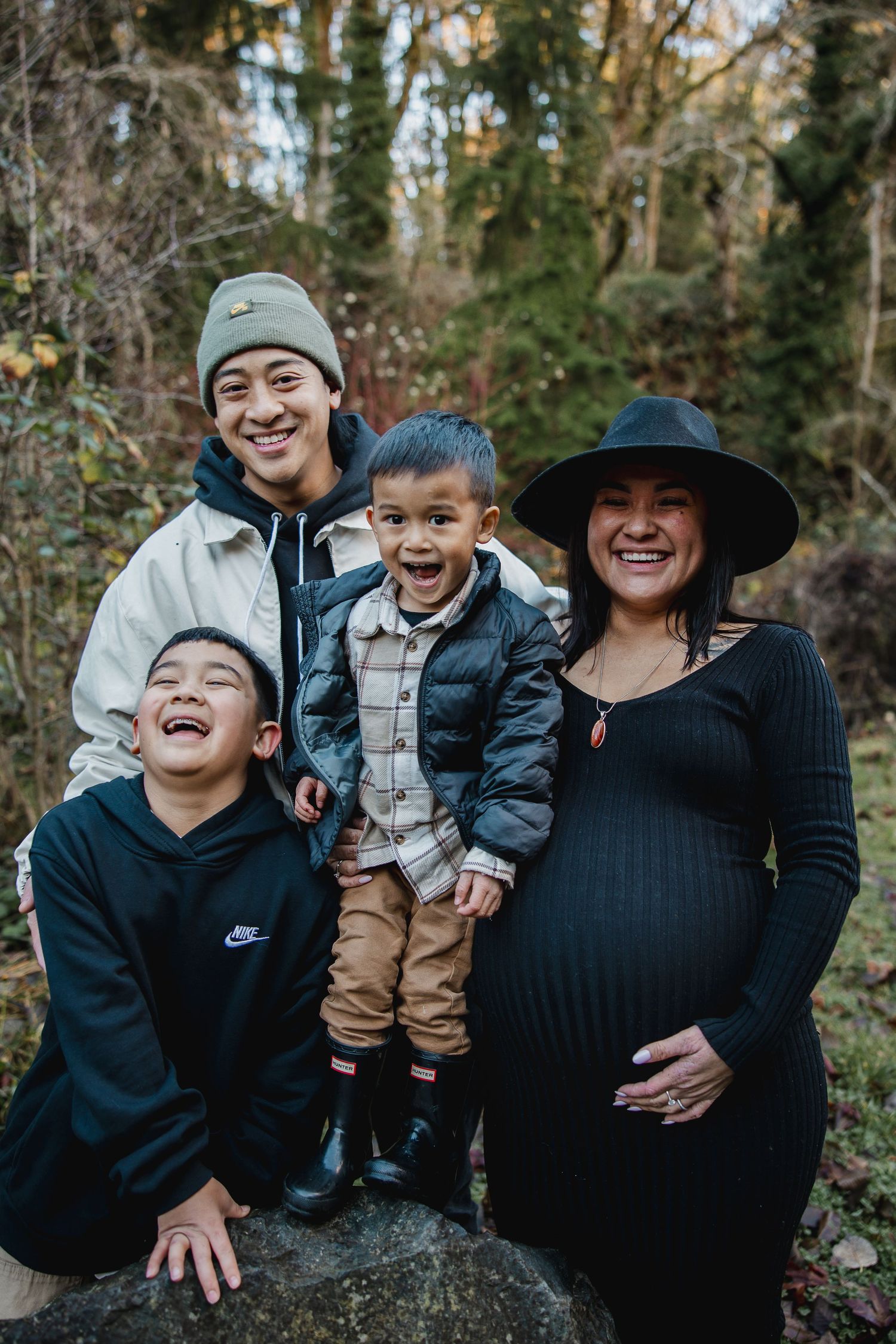 Pregnant woman in black dress and hat smiles with three boys outdoors in a lush forest setting.