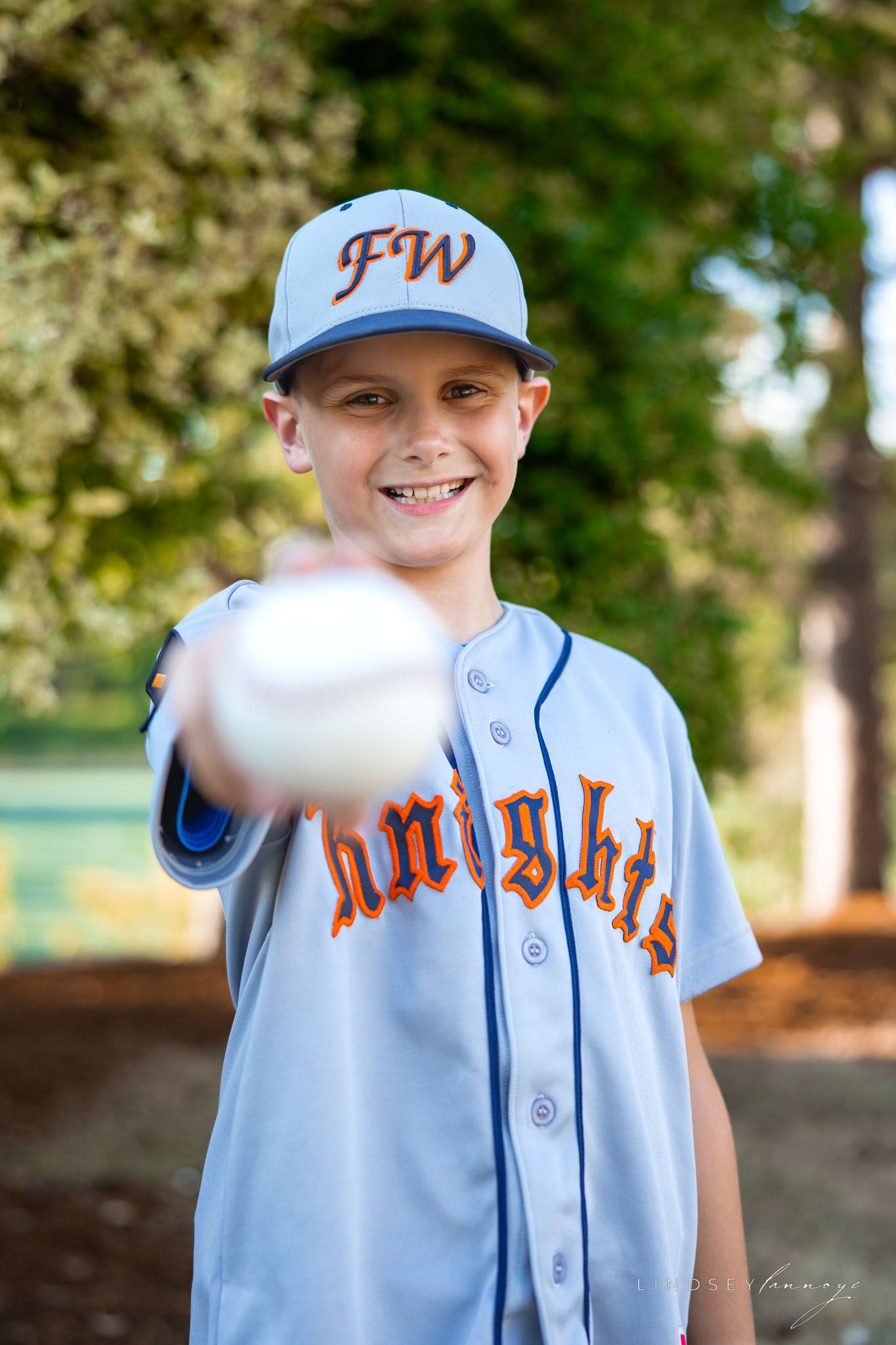 Young Knights baseball player in gray uniform smiles while holding a baseball toward the camera outdoors.