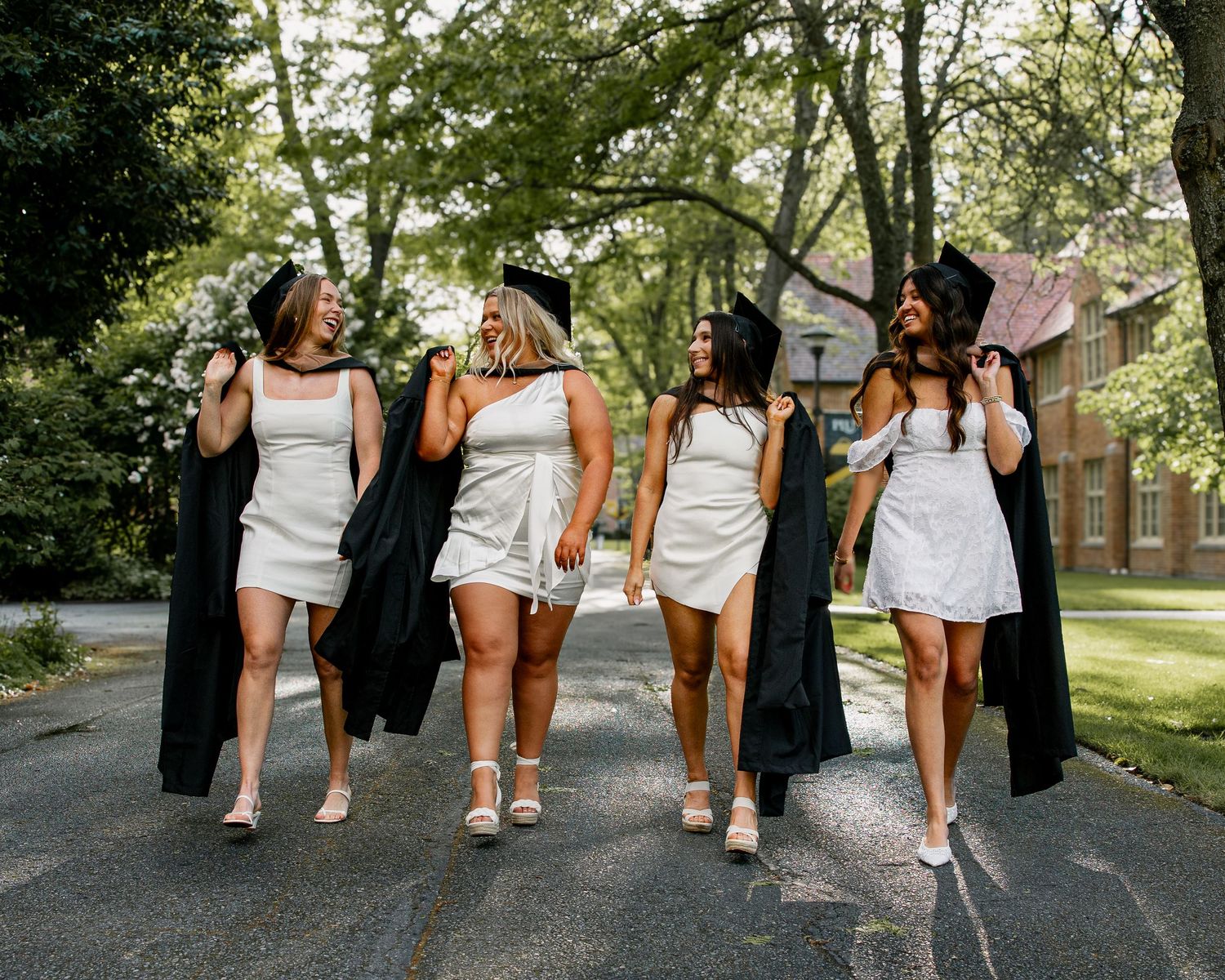 Five female graduates in white dresses carrying black graduation robes walking on sunny campus path.