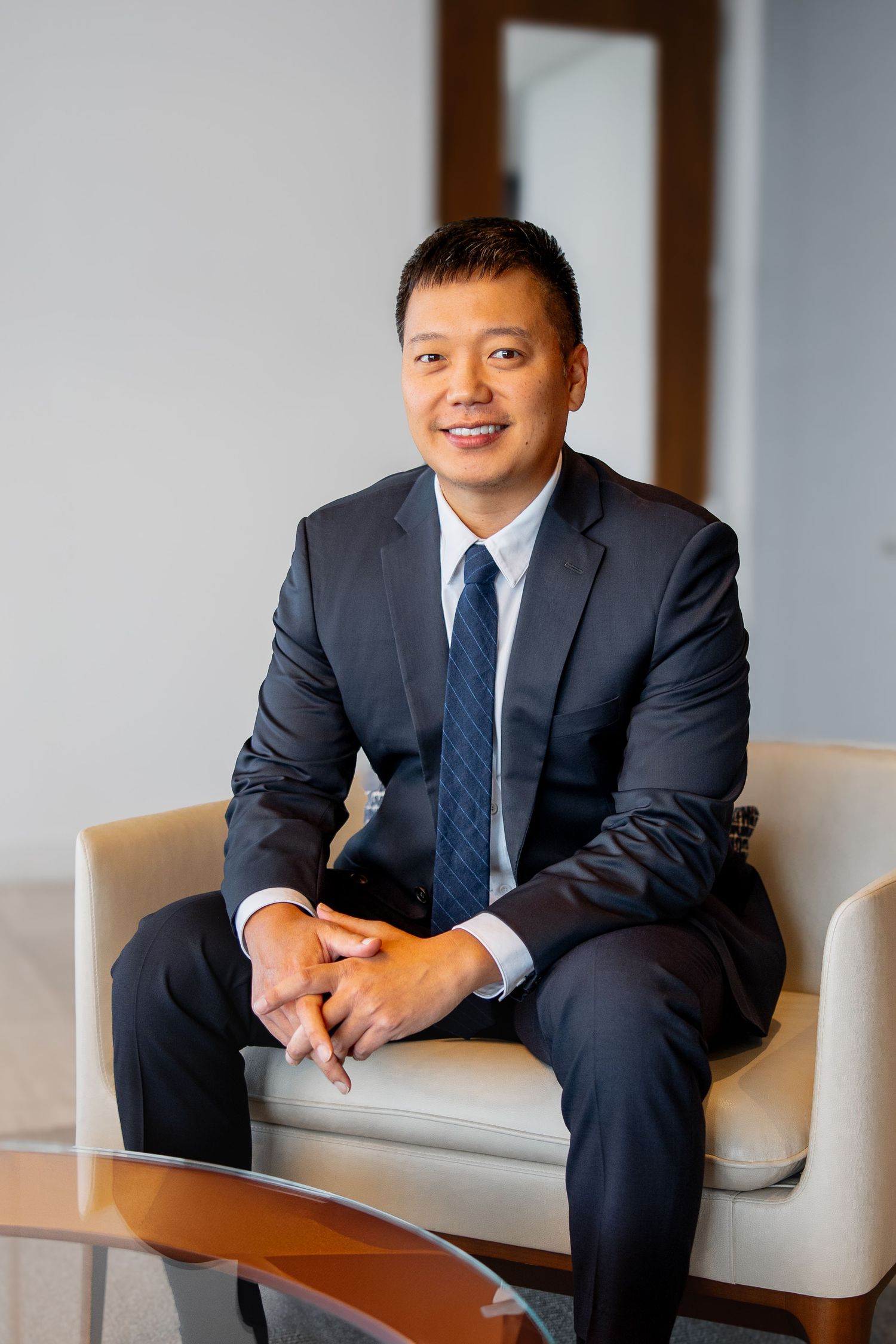 Smiling Asian businessman in navy suit seated in modern office lobby with clasped hands.