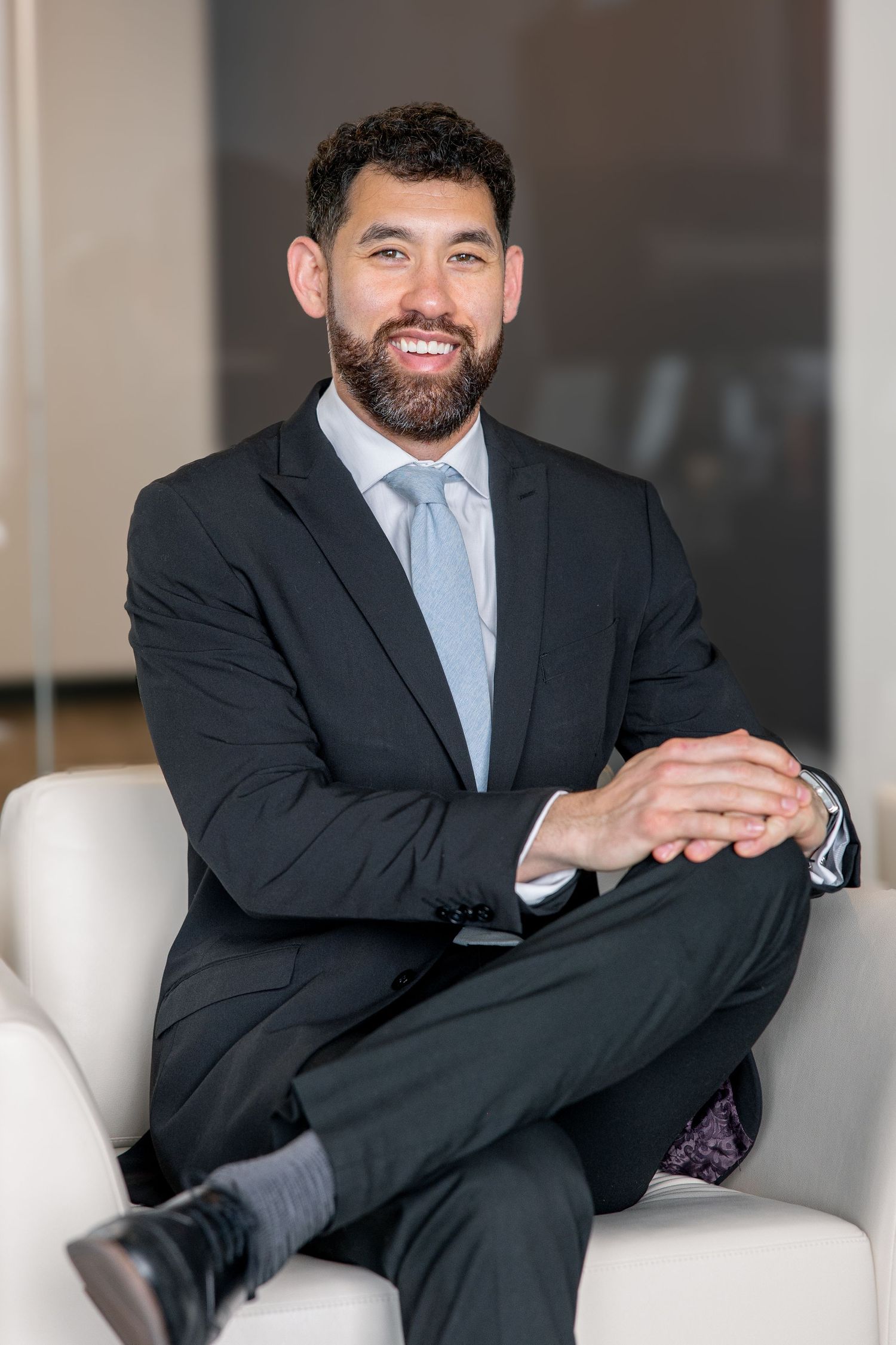 Confident bearded businessman in dark suit and tie seated with crossed legs in modern office.