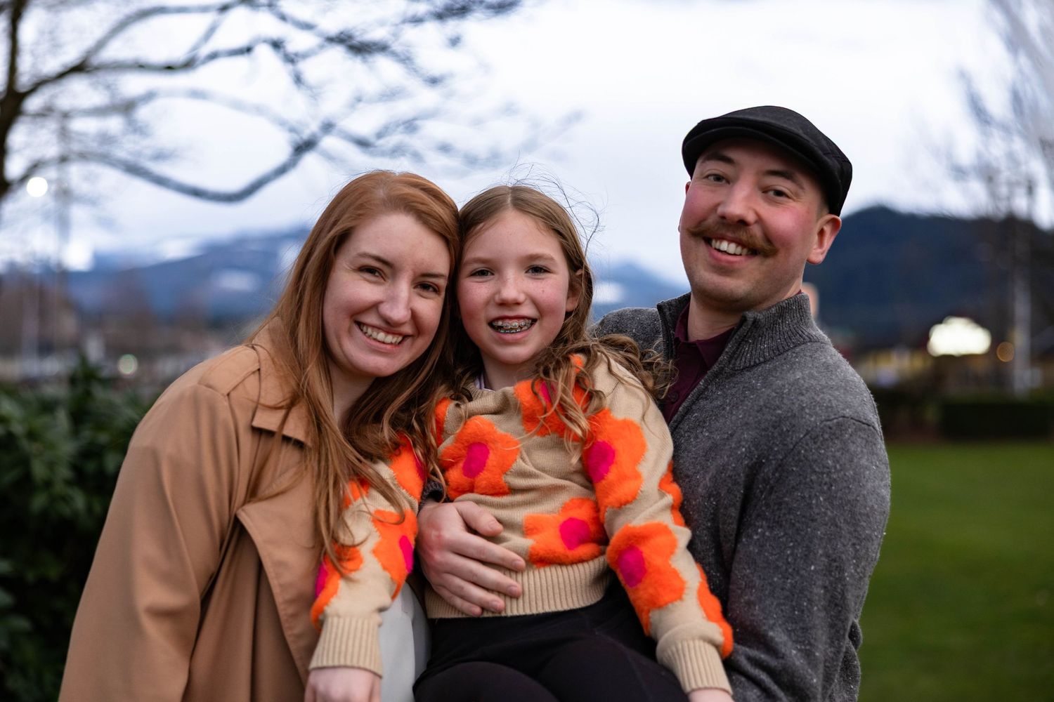 Smiling family of three posing outdoors with mountains and cloudy sky in background.