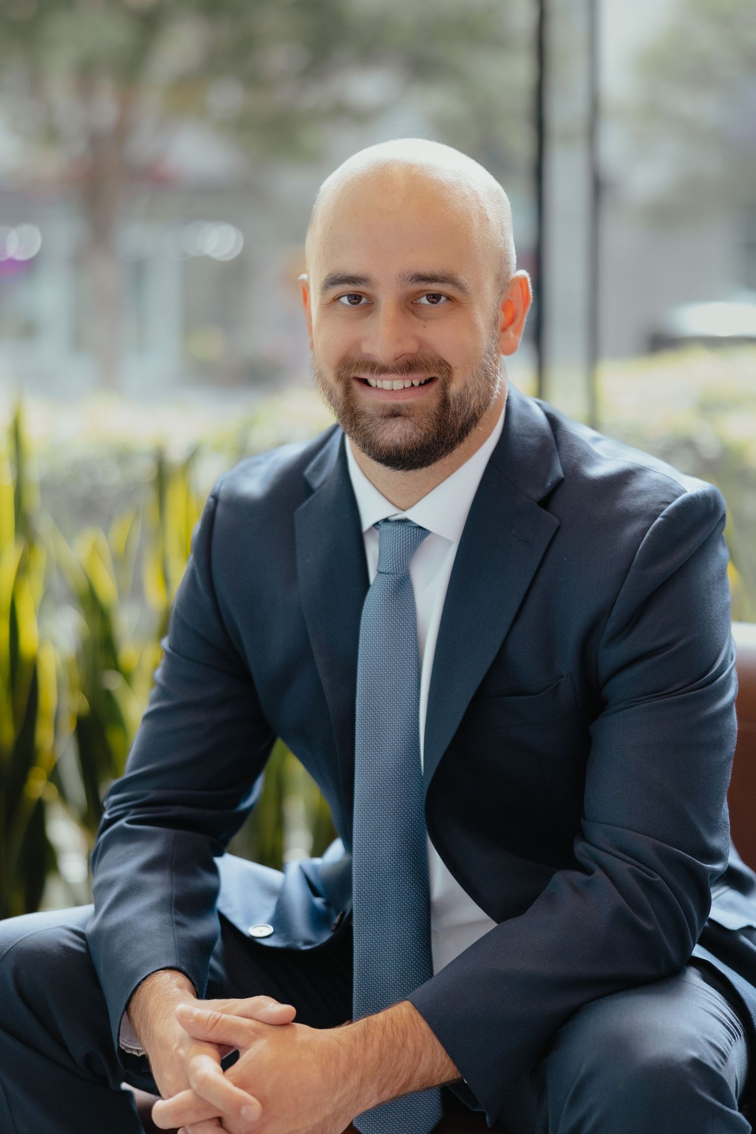 Smiling bald bearded man in dark suit sitting with hands clasped in modern indoor setting.