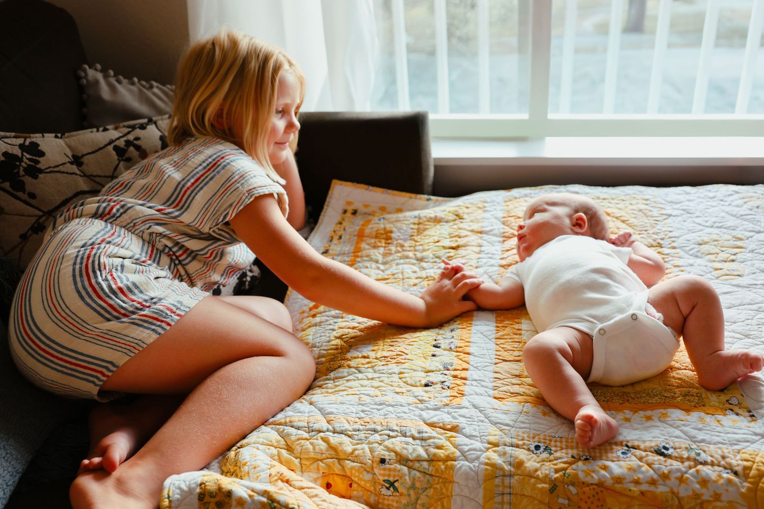 Young blonde girl sitting on bed smiling while gently touching newborn baby on yellow quilt.