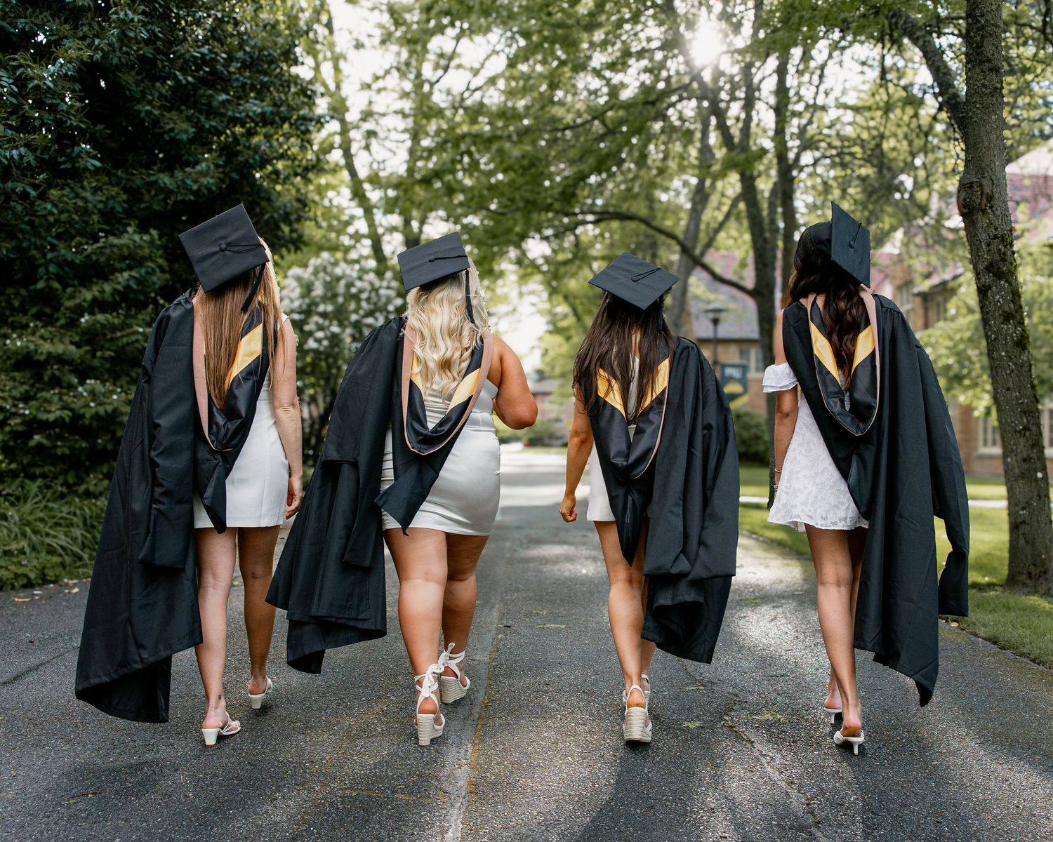 Four female graduates in black caps and gowns walking away down tree-lined campus pathway.