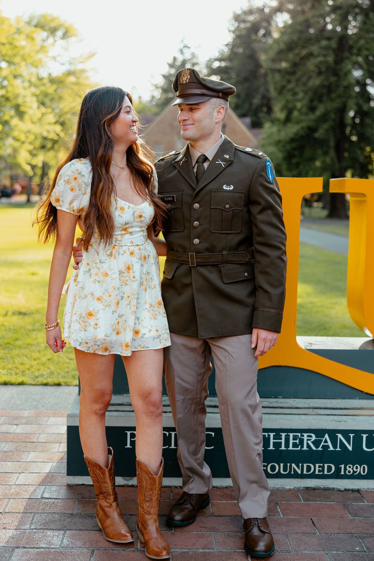 Couple posing outdoors, man in military uniform and woman in floral dress with cowboy boots, near a university sign.