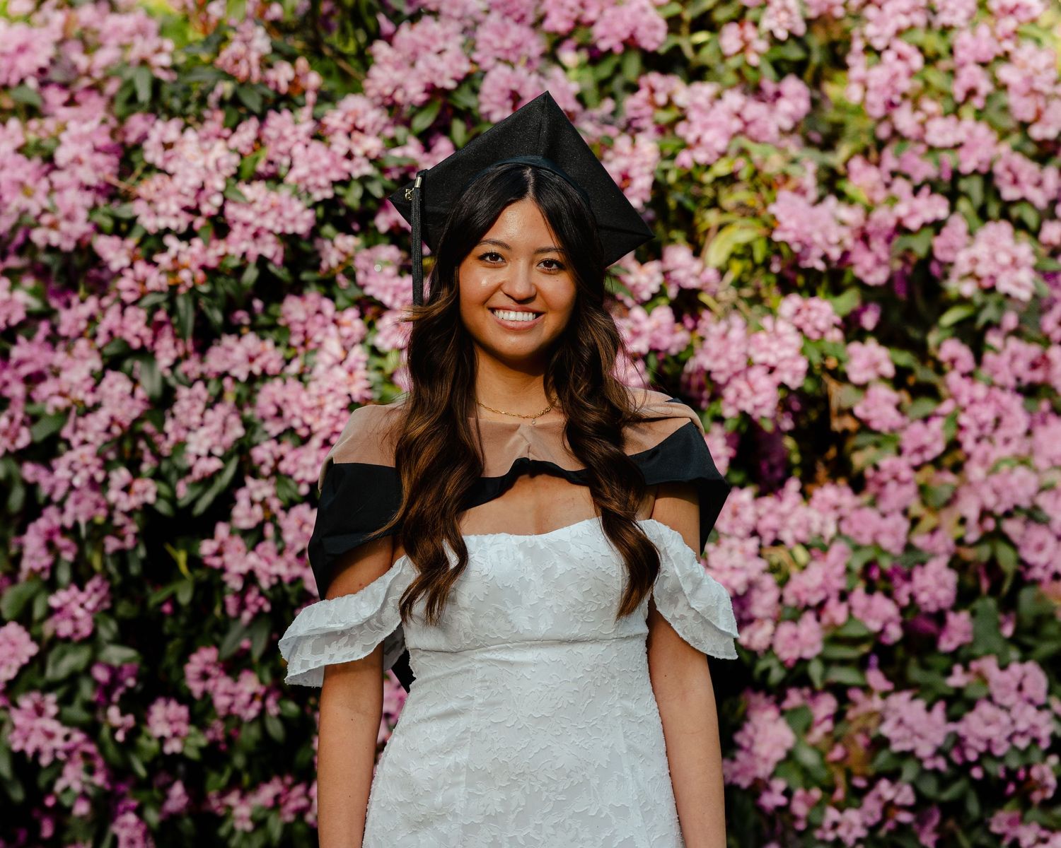 Smiling female graduate in white off-shoulder dress and black cap poses in front of vibrant pink flowering bushes.