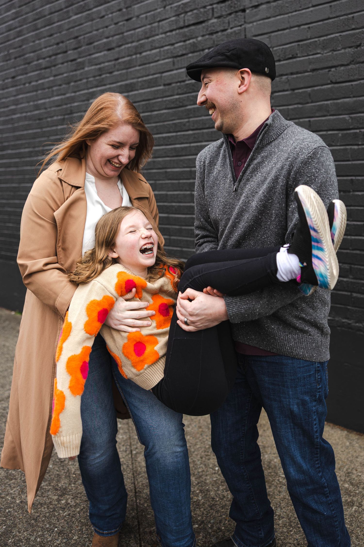 Joyful family with father lifting laughing girl in floral sweater against black brick wall.