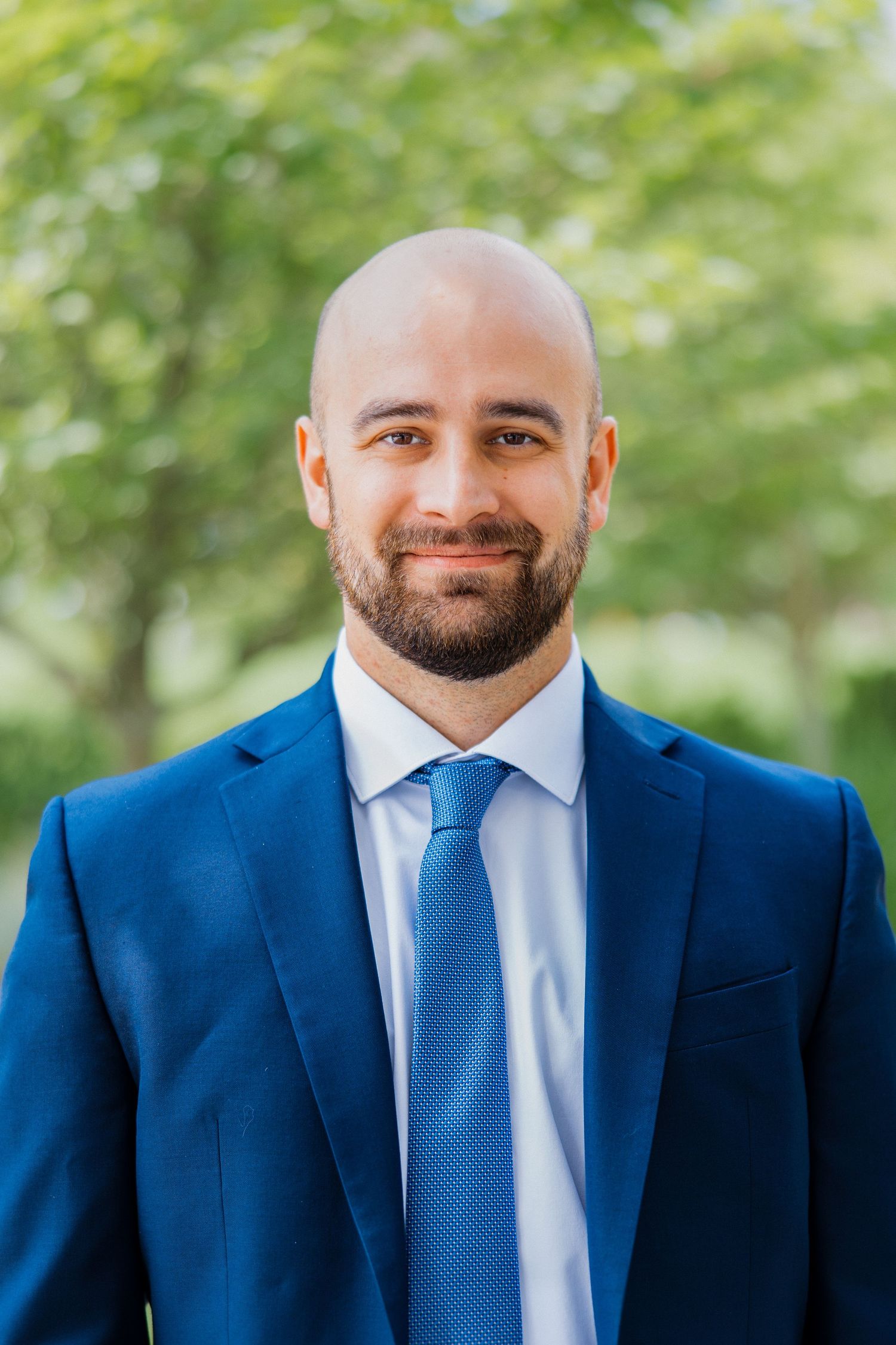 Bald man with beard in blue suit and tie smiling outdoors with green trees in background.