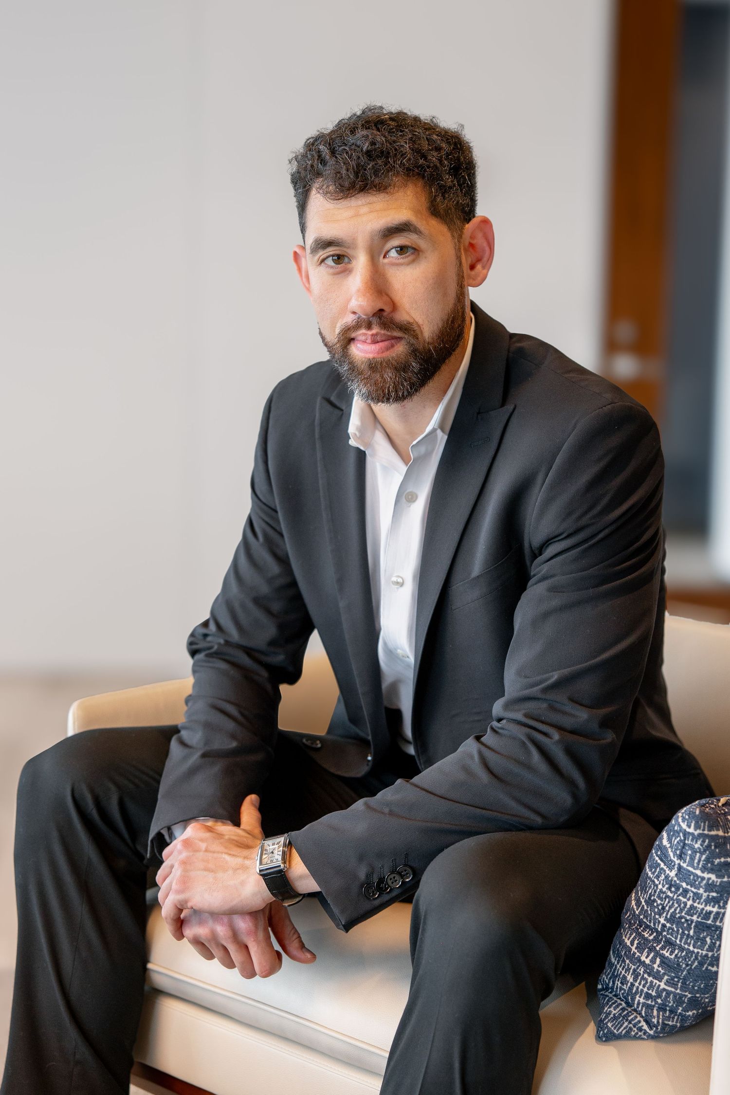 Professional man in dark suit seated in modern office lobby with serious expression.