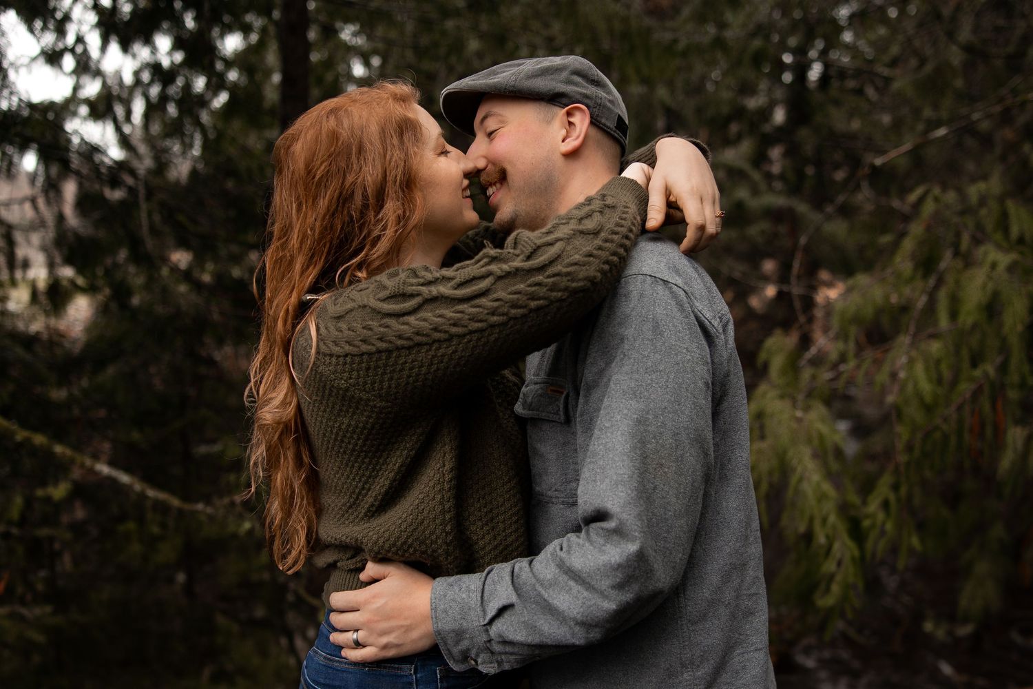 Couple sharing a warm embrace and nearly kissing outdoors among tall evergreen trees.