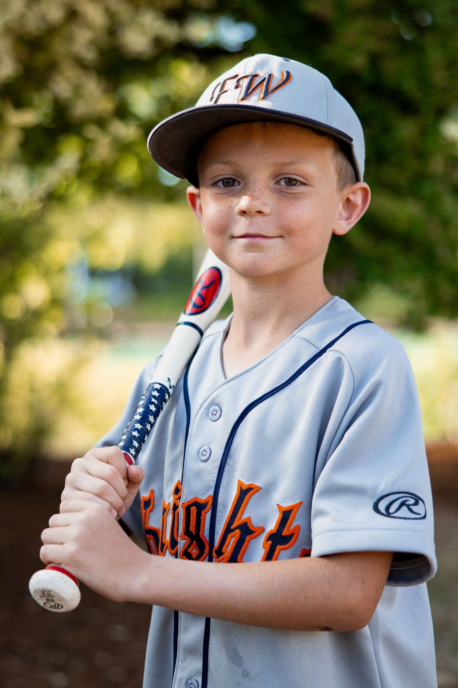 Smiling young boy in gray FW Knights baseball uniform holds a bat for a portrait outdoors.