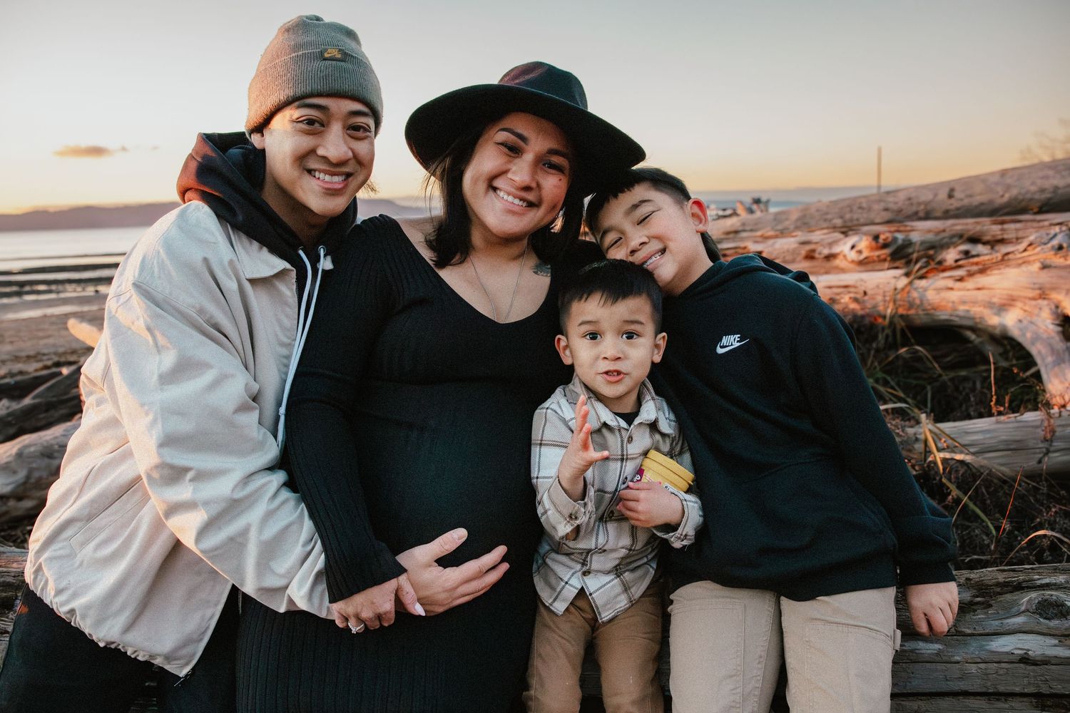 Pregnant woman in black dress and hat posing with family on a beach at sunset.